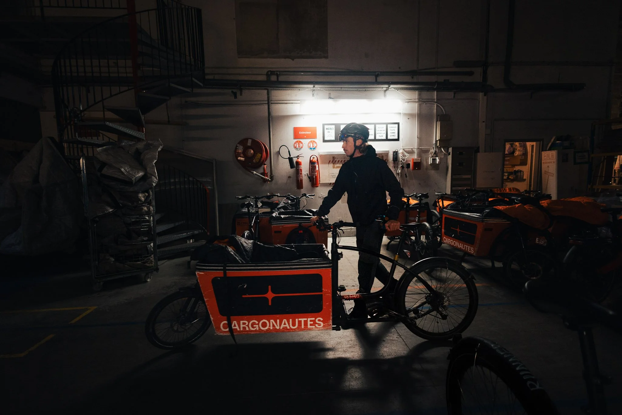 Person wearing a helmet standing next to an orange cargo bike marked with the words 'CARGONAUTES' in a dimly lit indoor parking area with other bikes and equipment.