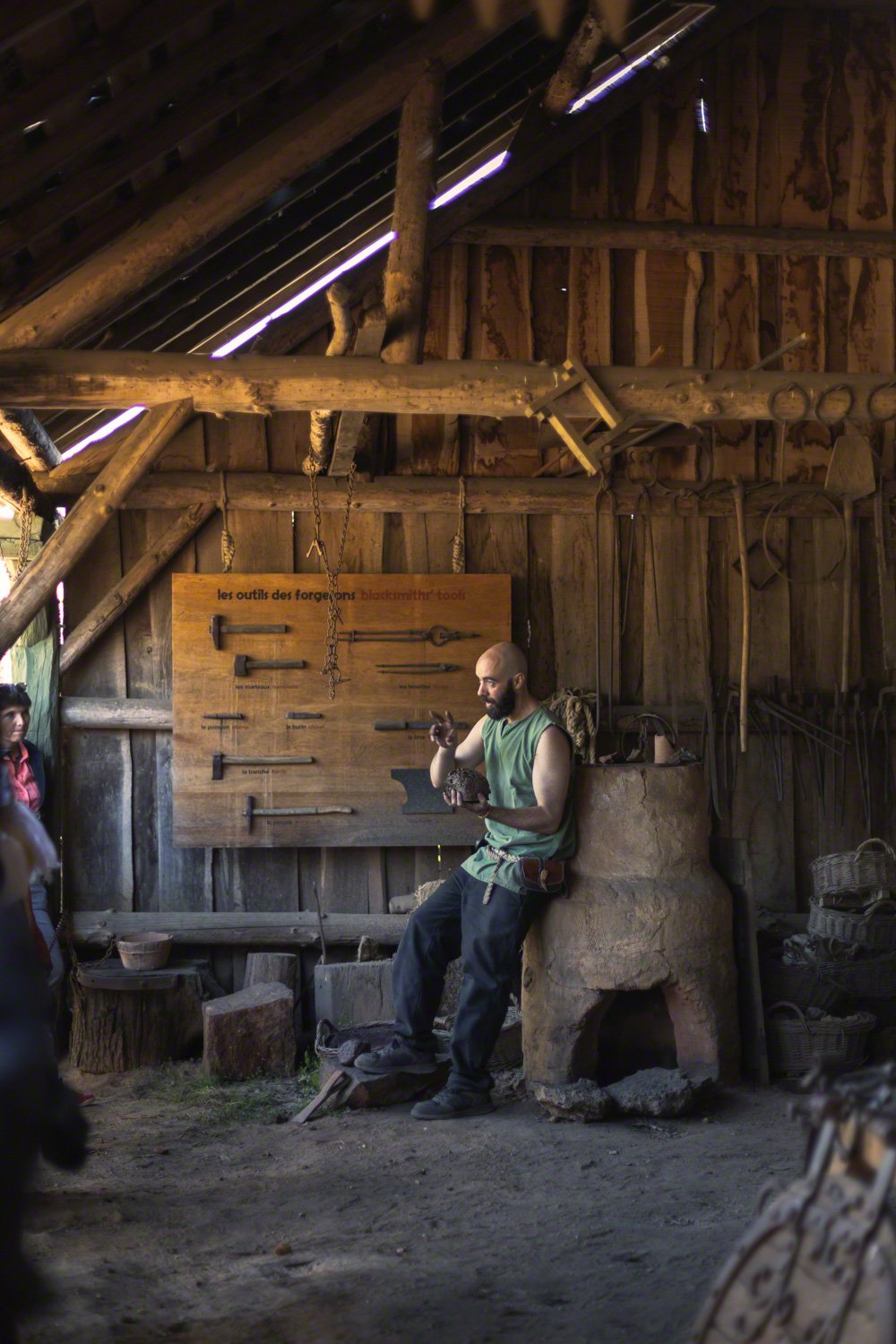 A man with a beard, wearing a green sleeveless shirt, is sitting on a stone structure inside a rustic workshop. He is holding an object and appears to be explaining or demonstrating something. Behind him, there is a wooden board with various smithing