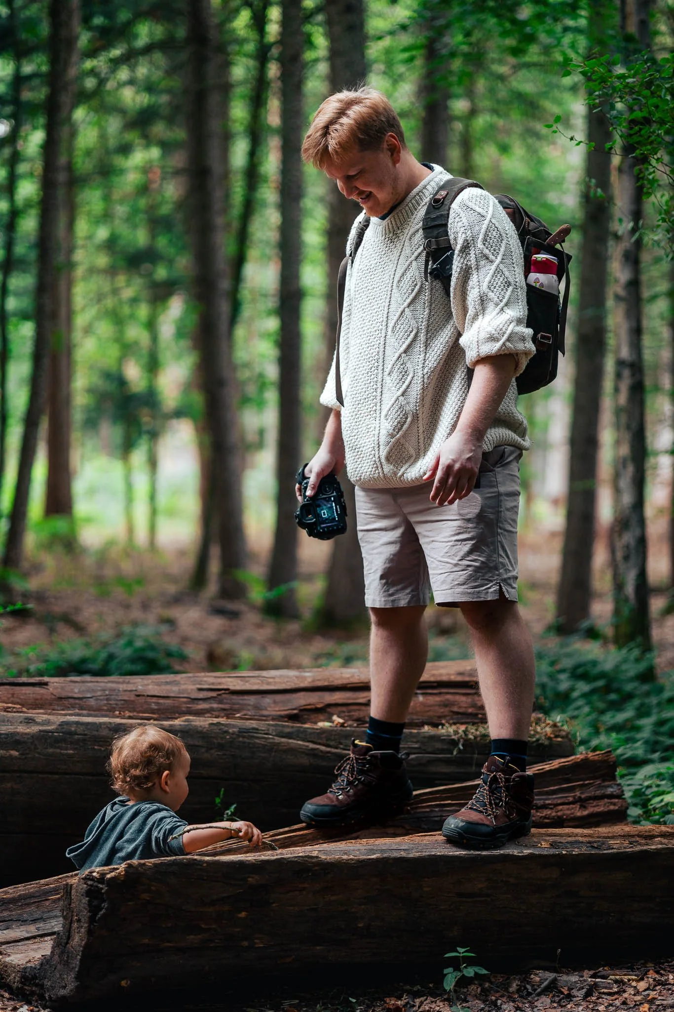 A young man with a camera and backpack hiking in a wooded forest, looking at a small child climbing on a fallen log.