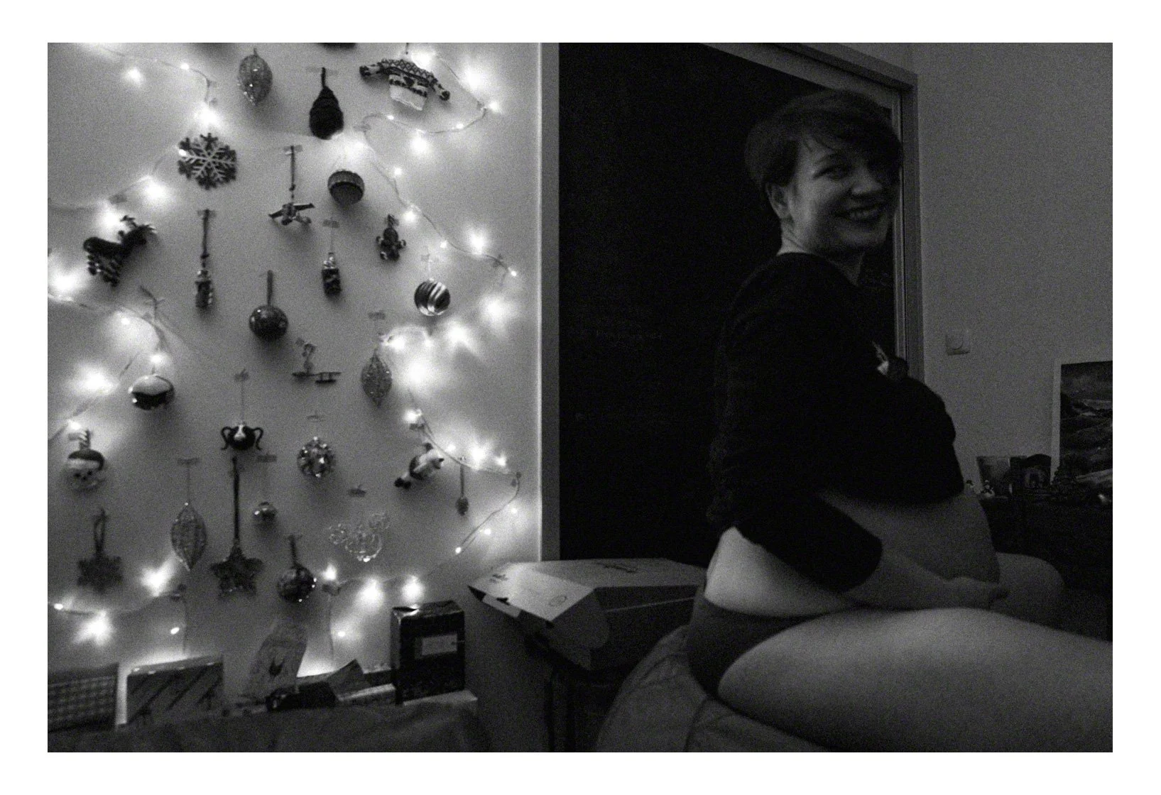 A woman smiling on her stomach, with her arms crossed in a cozy room decorated for Christmas, featuring a tree with ornaments and lights on the wall behind her.