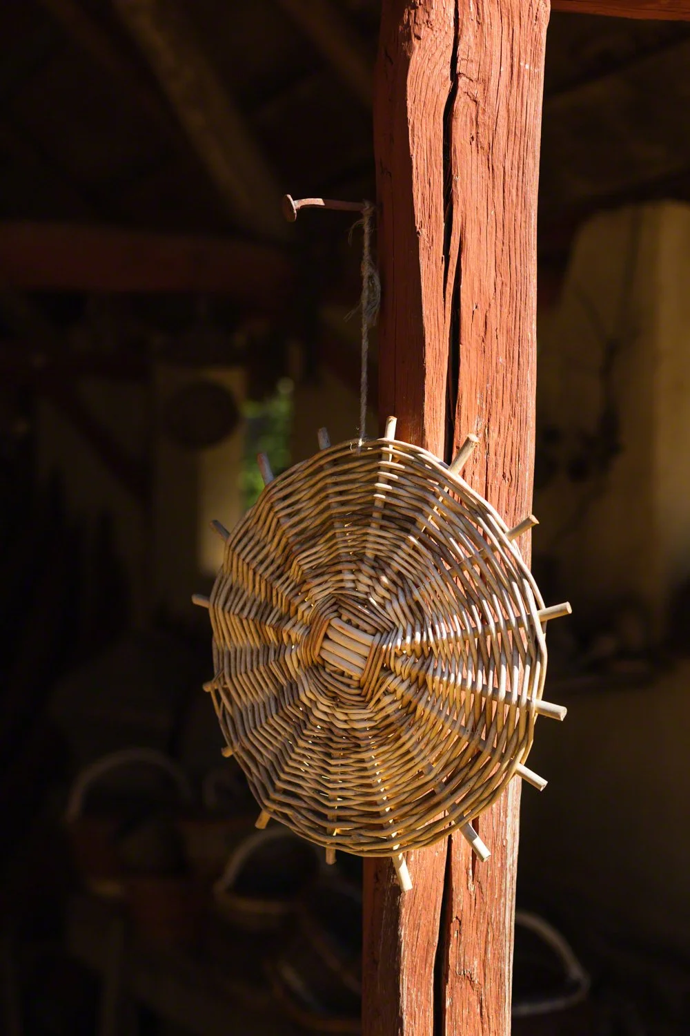 A woven basket with wooden sticks protruding from its edges, hanging on a worn, red-painted wooden beam in a rustic setting.