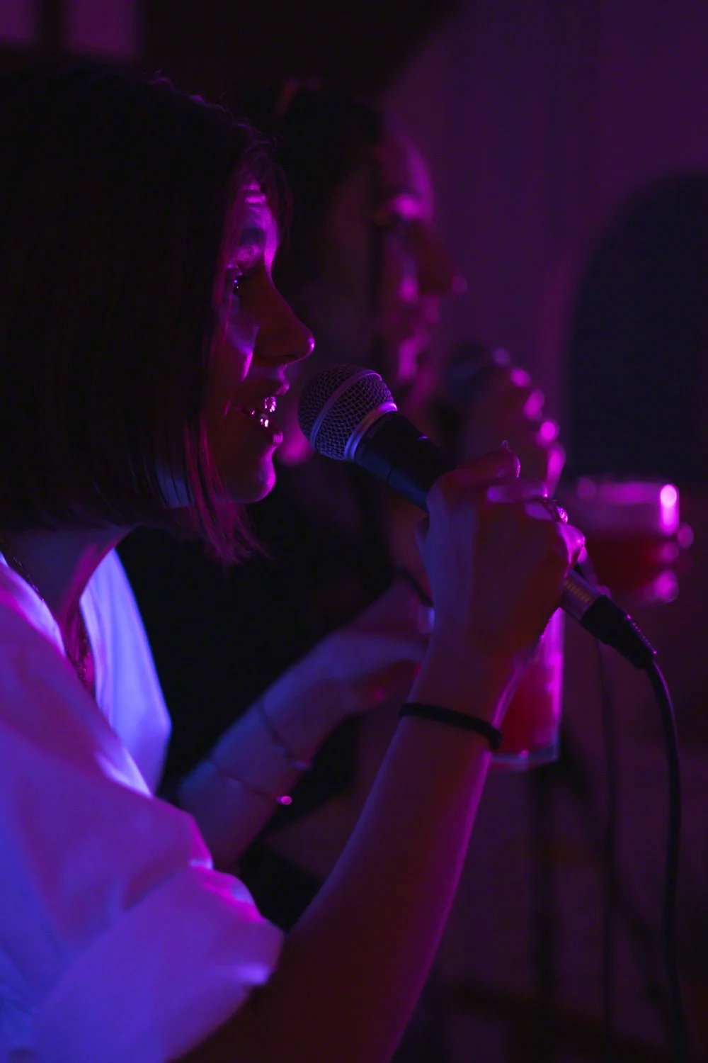 A woman singing into a microphone in a dimly lit room with purple lighting, with other people also singing into microphones in the background.