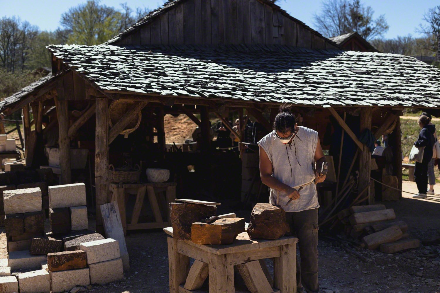 A person working with pottery under a wooden, rustic shelter on a sunny day.