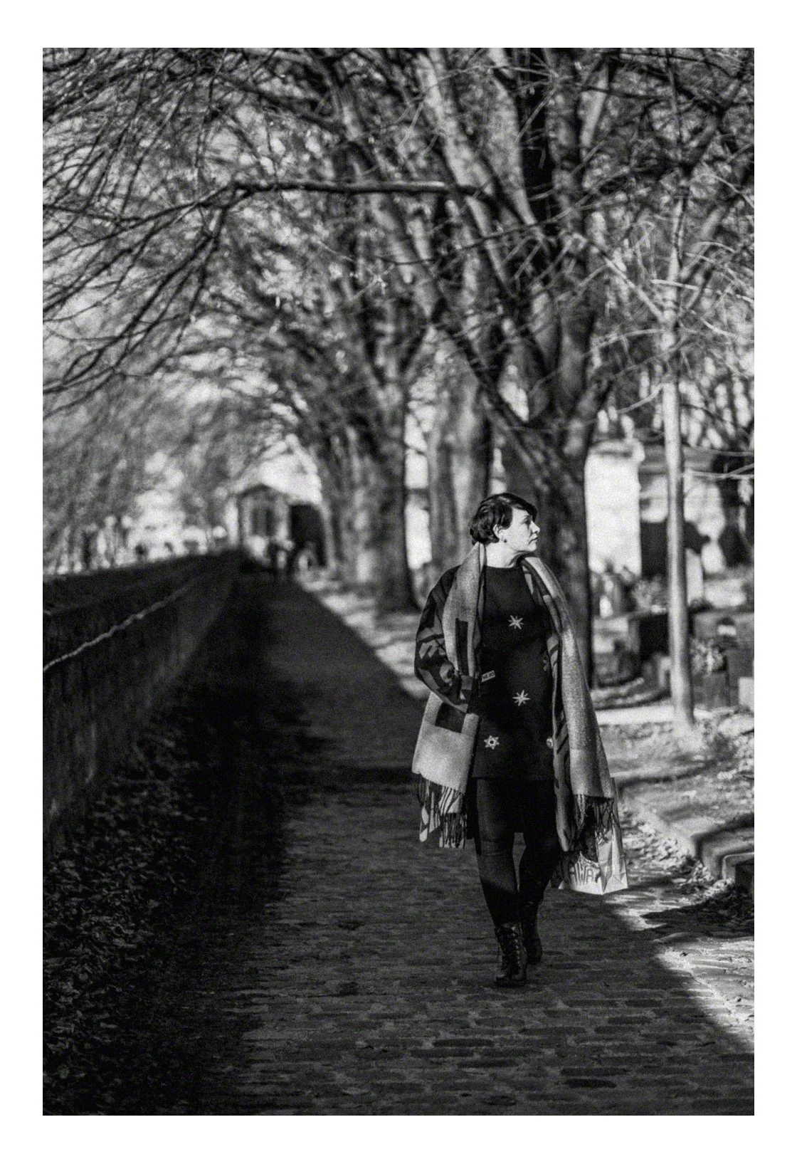 A woman walking on a cobblestone sidewalk lined with leafless trees on a winter day, wearing a patterned coat and scarf, in black and white.