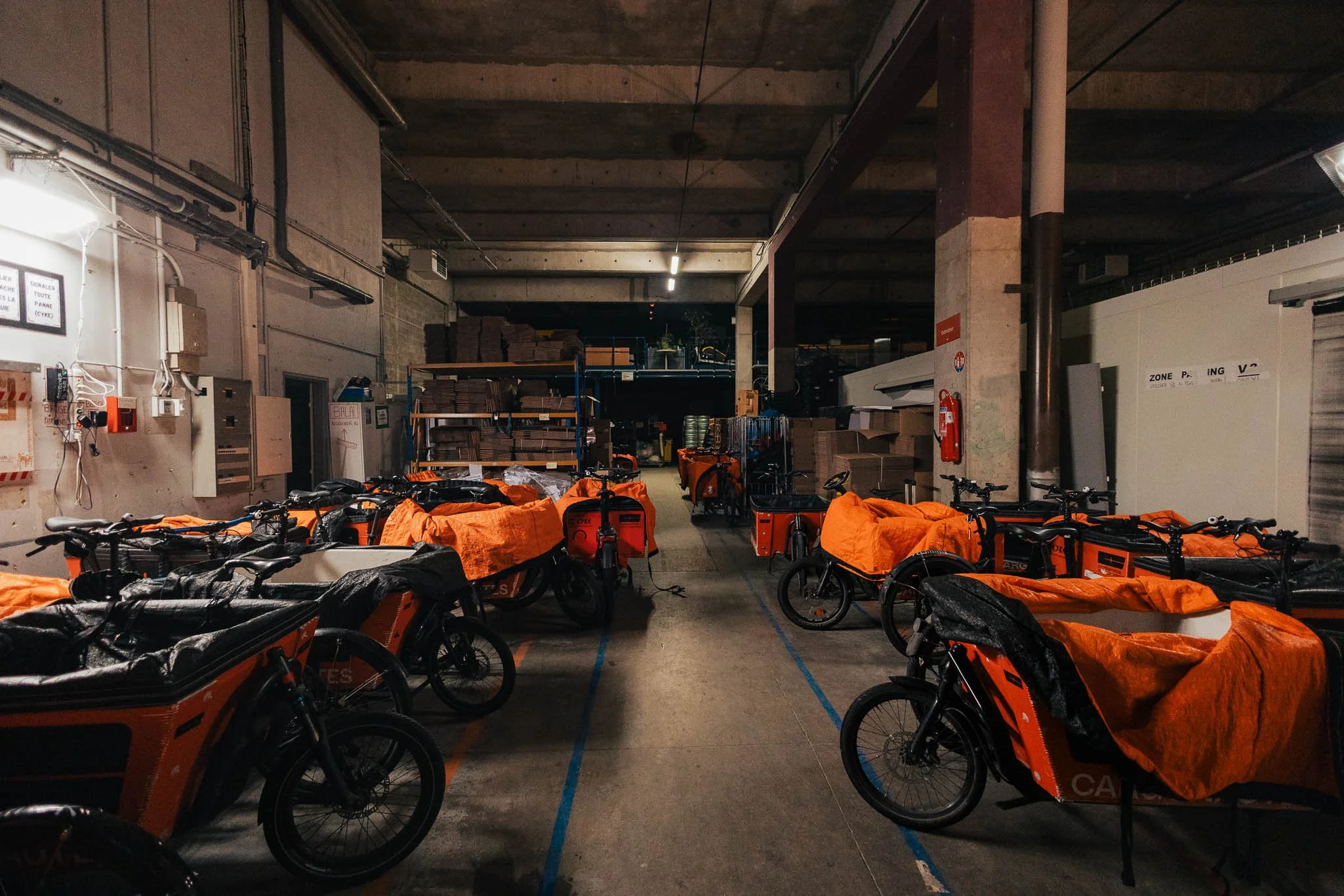 Indoor warehouse or storage area with multiple orange delivery bicycles lined up, covered with orange tarps, and some shelves with boxes and supplies in the background.