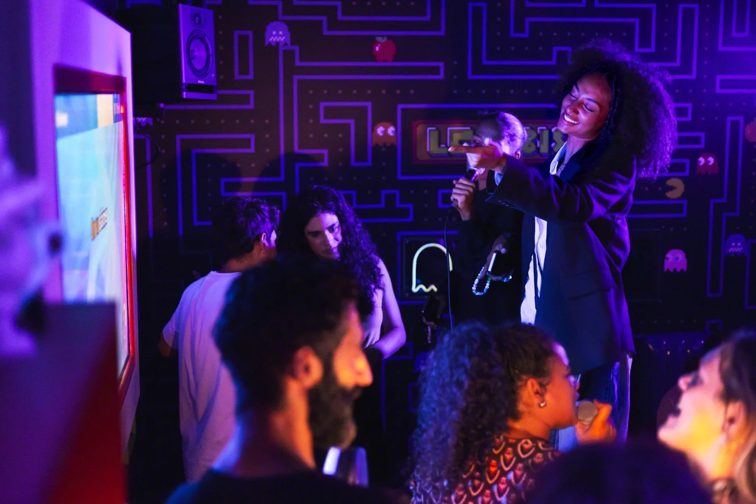 A woman with curly hair in a black blazer holding a microphone in front of a small group of people in a dimly lit arcade or game room featuring pixel art from classic video games.