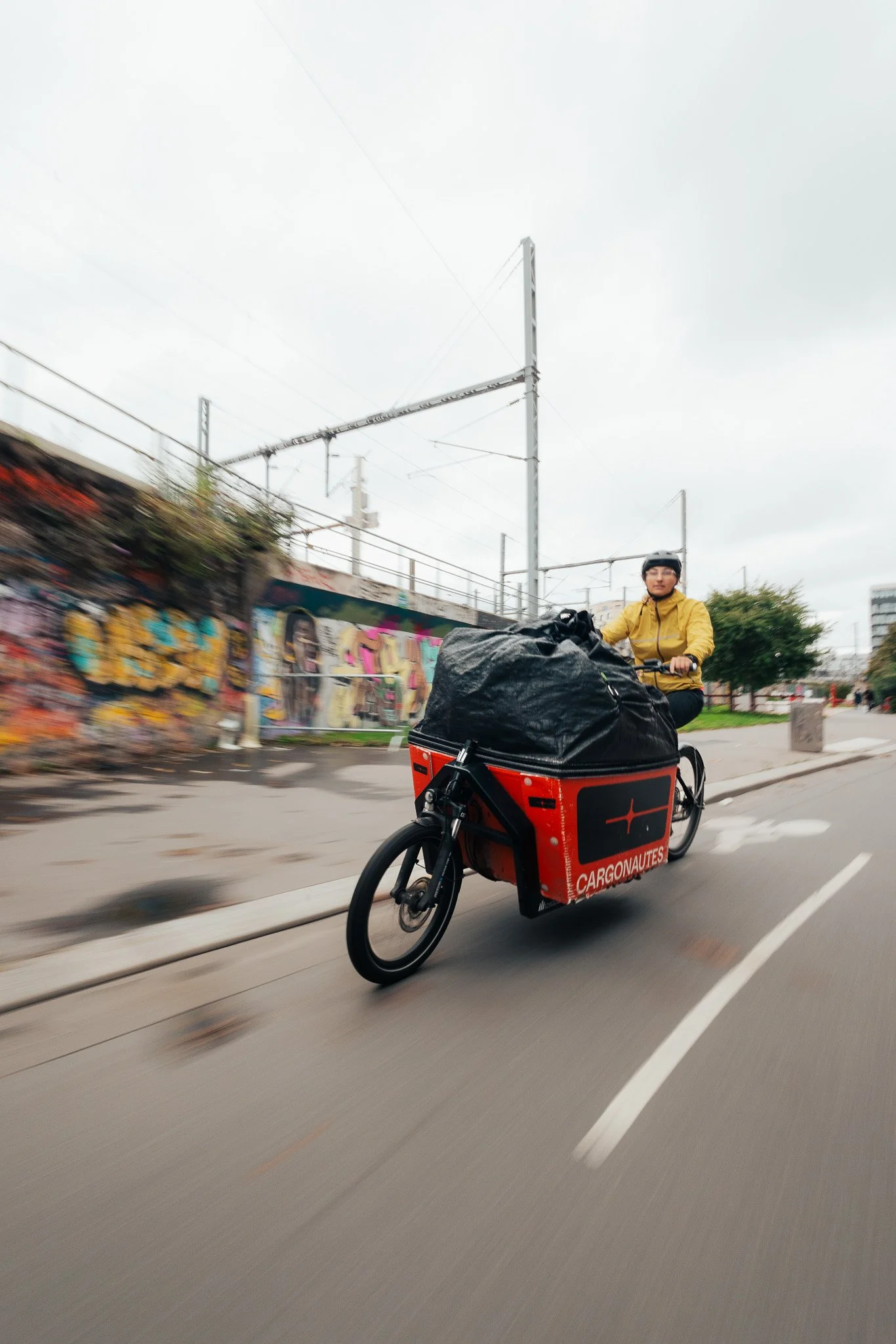 A person riding a cargo e-bike on a city street, wearing a yellow jacket and helmet, with a large black bag loaded on the bike, graffiti on the wall in the background.