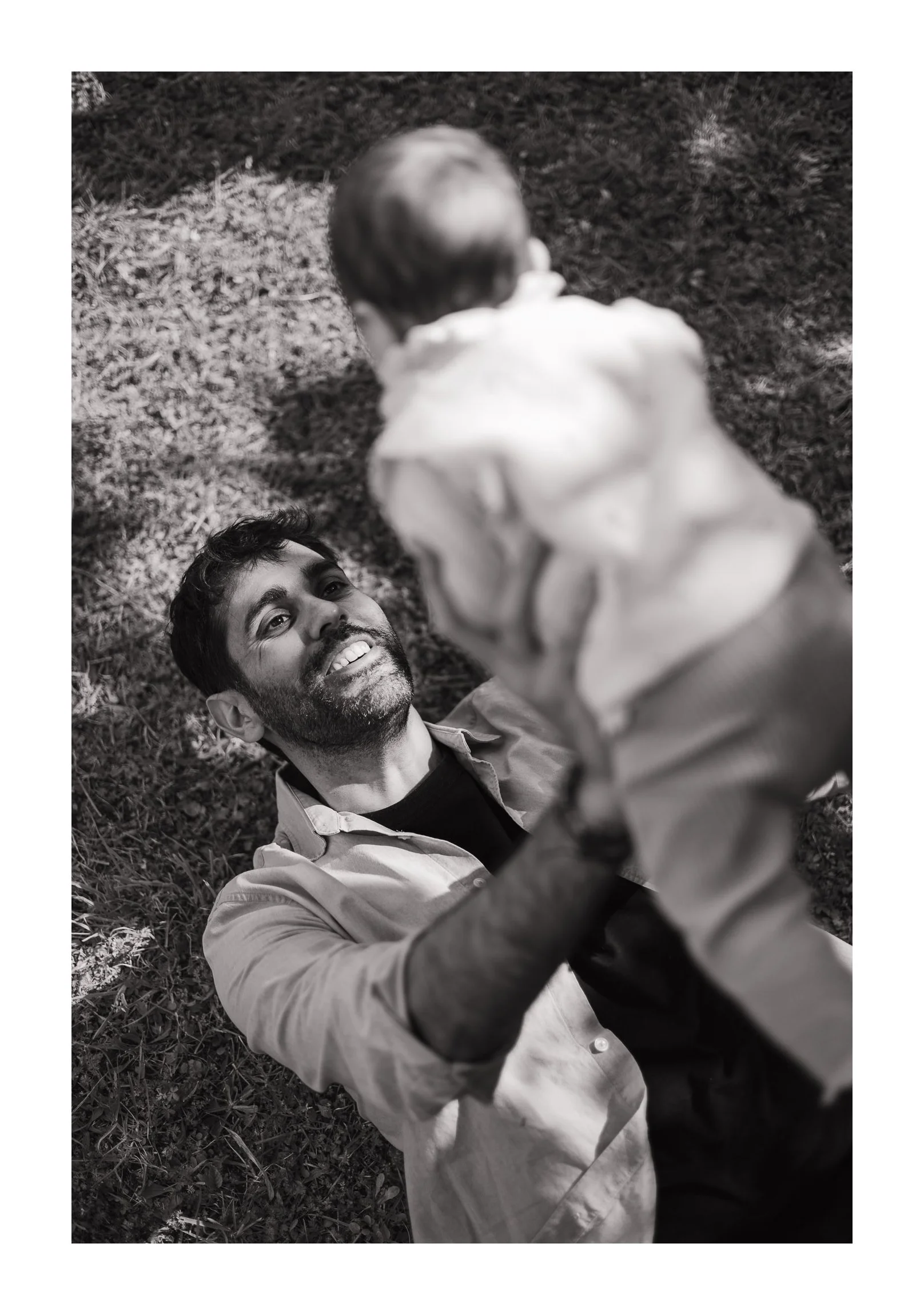 A man lying on the grass, smiling, holding a small child above him, seen from above, in black and white.