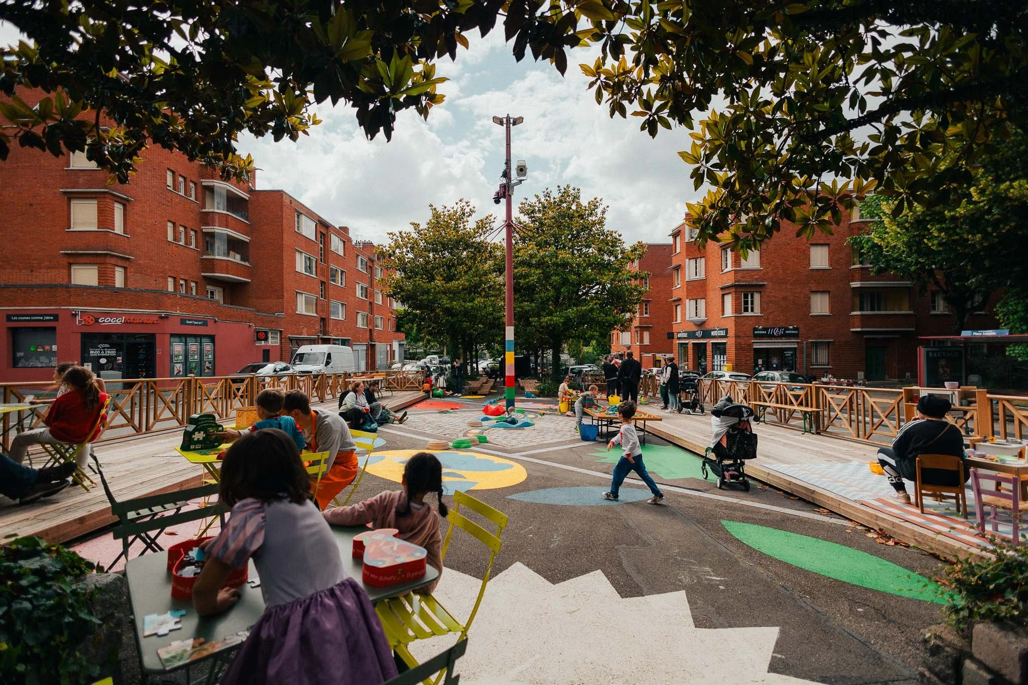 An outdoor playground with children and adults, surrounded by red brick apartment buildings, trees, and a cloudy sky.