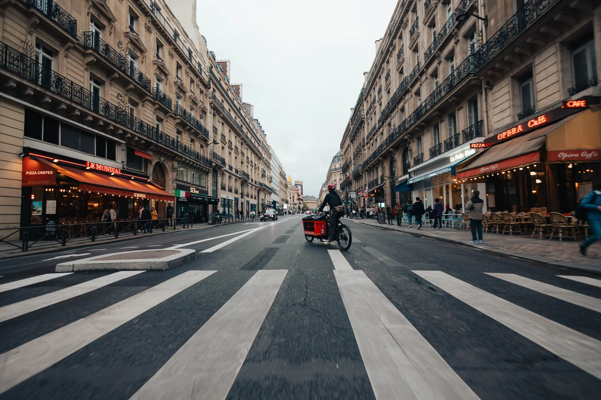 City street scene with buildings lining both sides, a cyclist crossing at a crosswalk, pedestrians walking along sidewalks, and various storefronts, including a pizza restaurant and cafes.