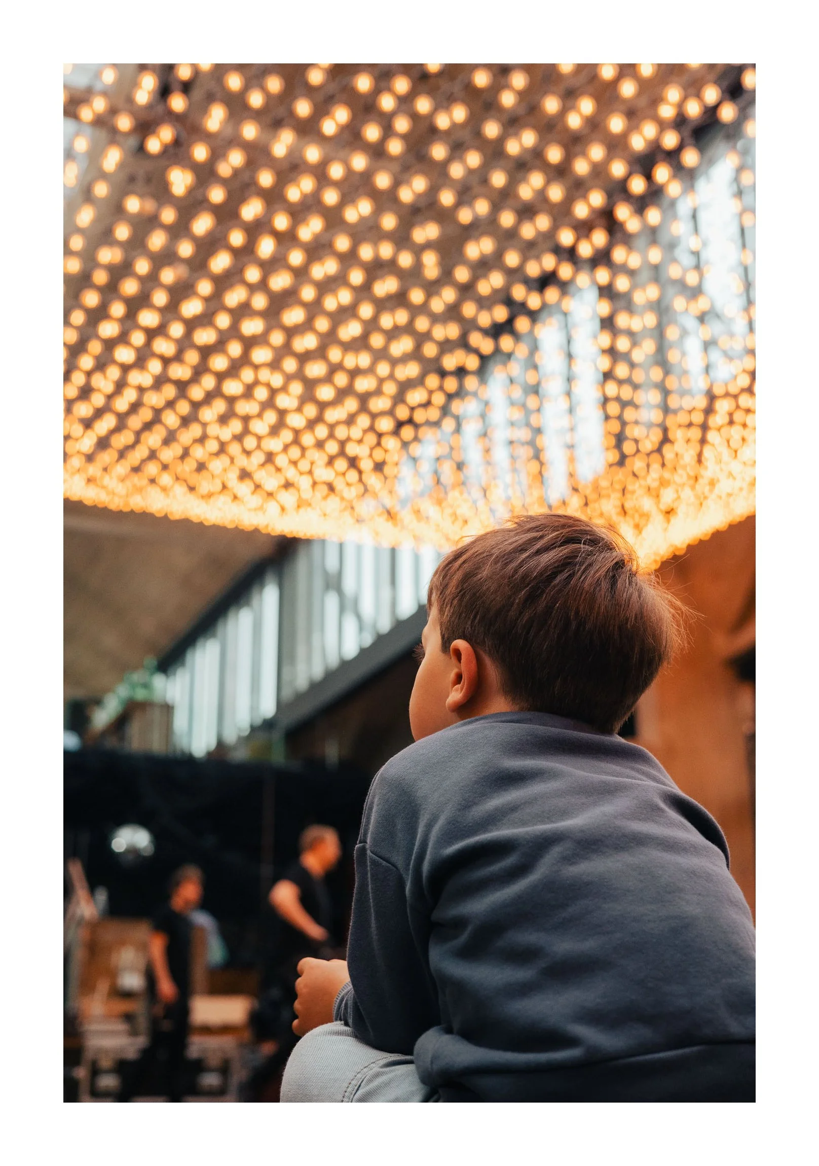 Child sitting indoors under a ceiling decorated with numerous warm-colored string lights, with a blurry background of other people and large windows.