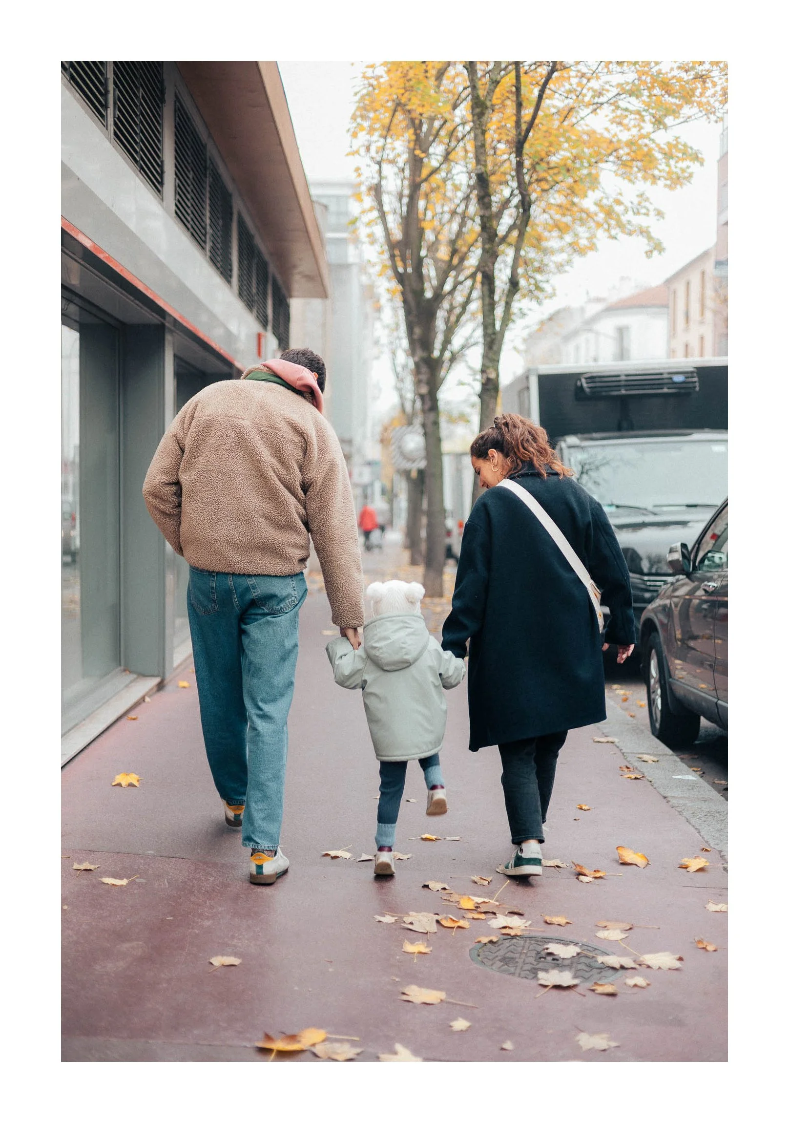 A family of three walking on a city sidewalk in autumn, holding hands, with fallen leaves on the ground and trees with yellow leaves in the background.