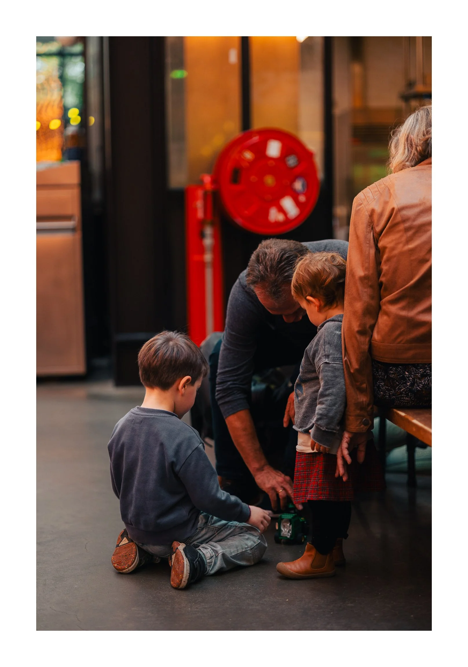 A man, woman, and two children playing with a toy car inside a room, with a red fire hose reel in the background.