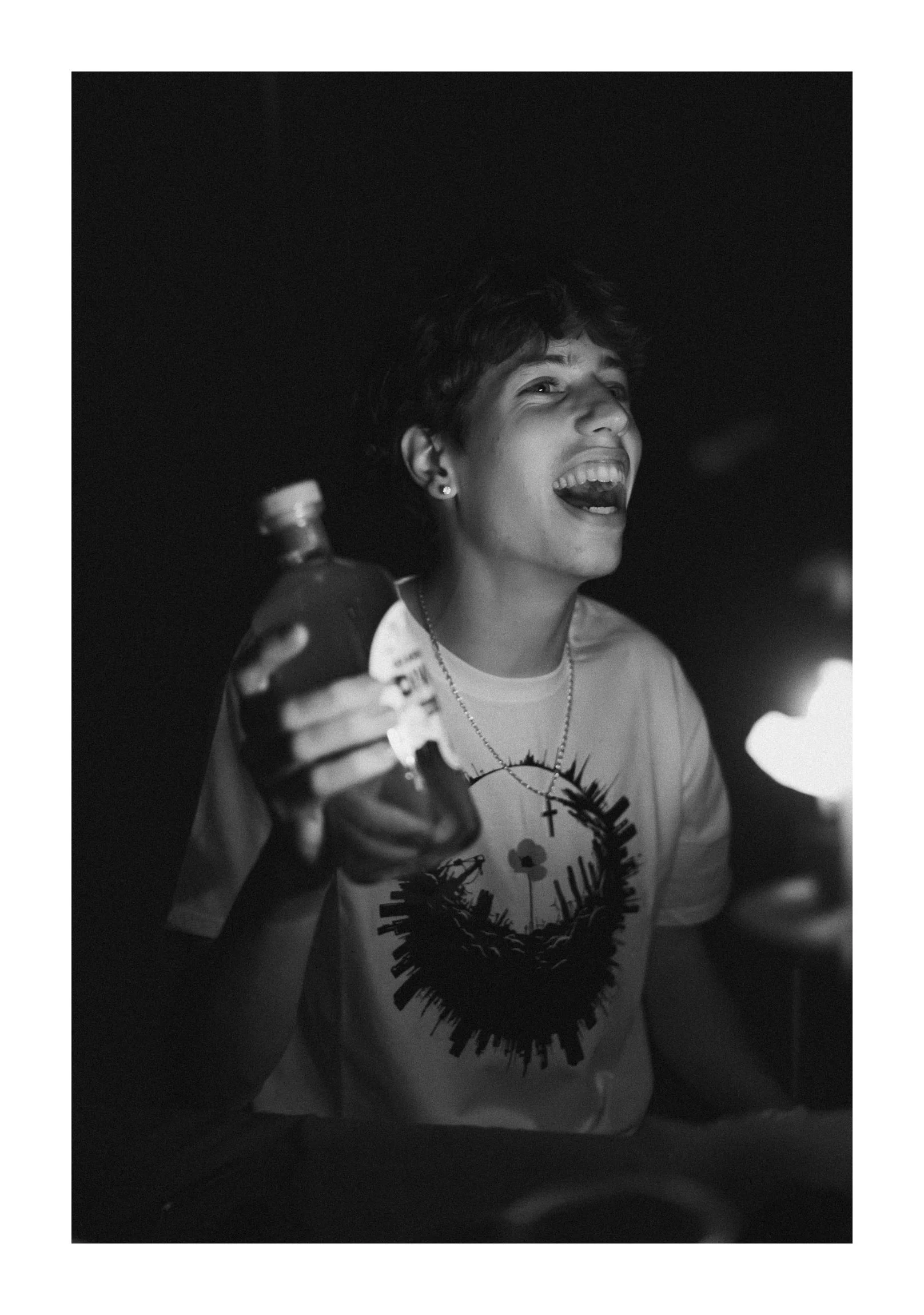 A person smiling and laughing, holding a bottle, in a dark setting with a light source on the right.