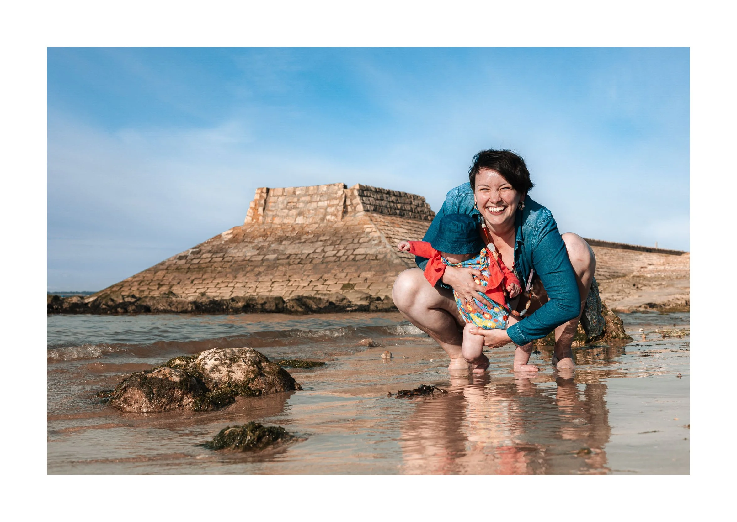 A woman and a child smiling and crouching on a beach with rocks and water, with an ancient stone fortress in the background and a clear blue sky.
