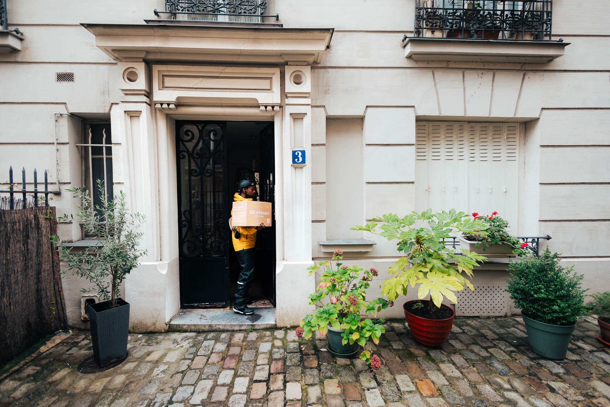 A delivery person wearing a yellow jacket and black helmet standing at a black gate, holding a brown box with white and orange text. Potted plants line a cobblestone sidewalk outside a beige building with the number 3 and small balconies with plants.