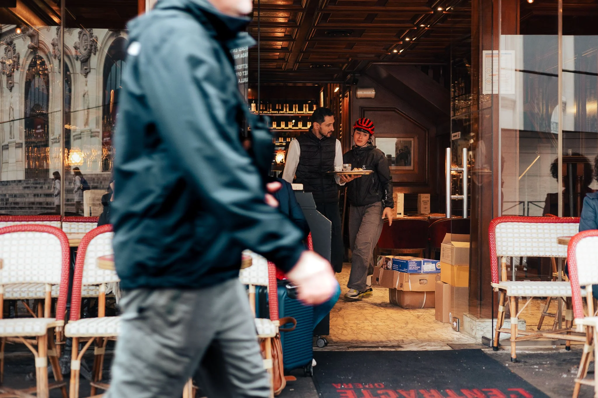 People entering a restaurant or cafe with a person in a black jacket and a woman with a red striped bicycle helmet inside.