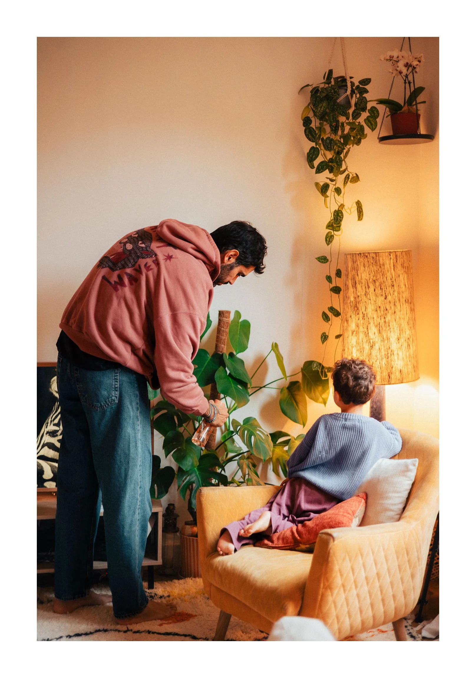 A man and a child in a living room with warm lighting, plants, and a sofa. The man is standing next to the child, who is sitting on the sofa facing away from the camera.
