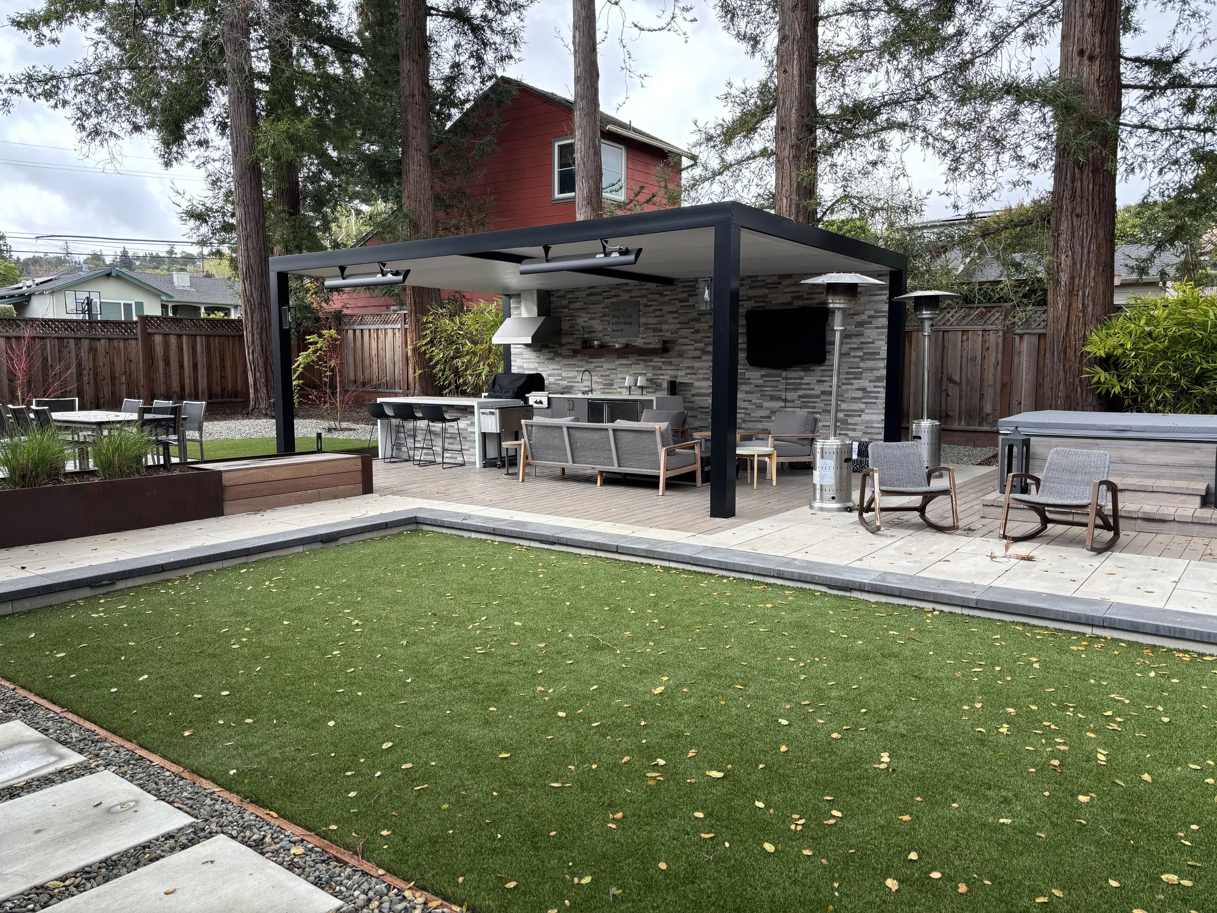 Modern backyard patio with a covered outdoor kitchen, bar seating, lounge furniture, heaters, artificial turf, and cedar trees