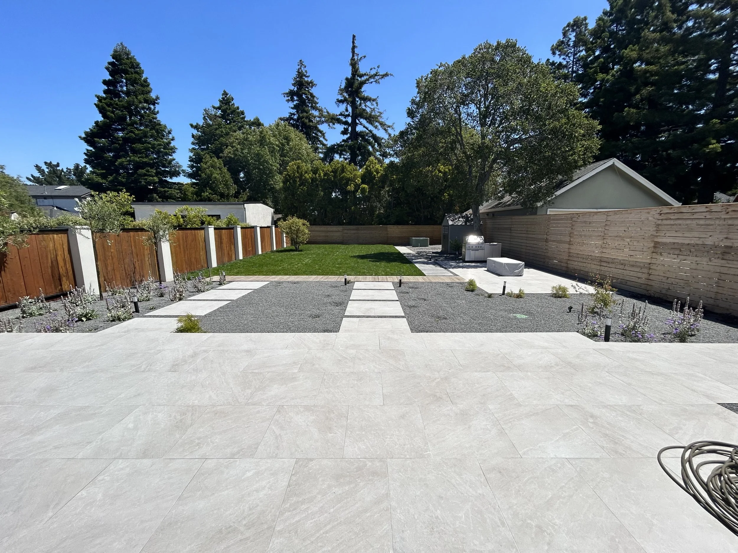 Modern backyard with large tiled patio, gravel landscaping, lawn, garden beds, wood fencing, outdoor grill, and mature trees.