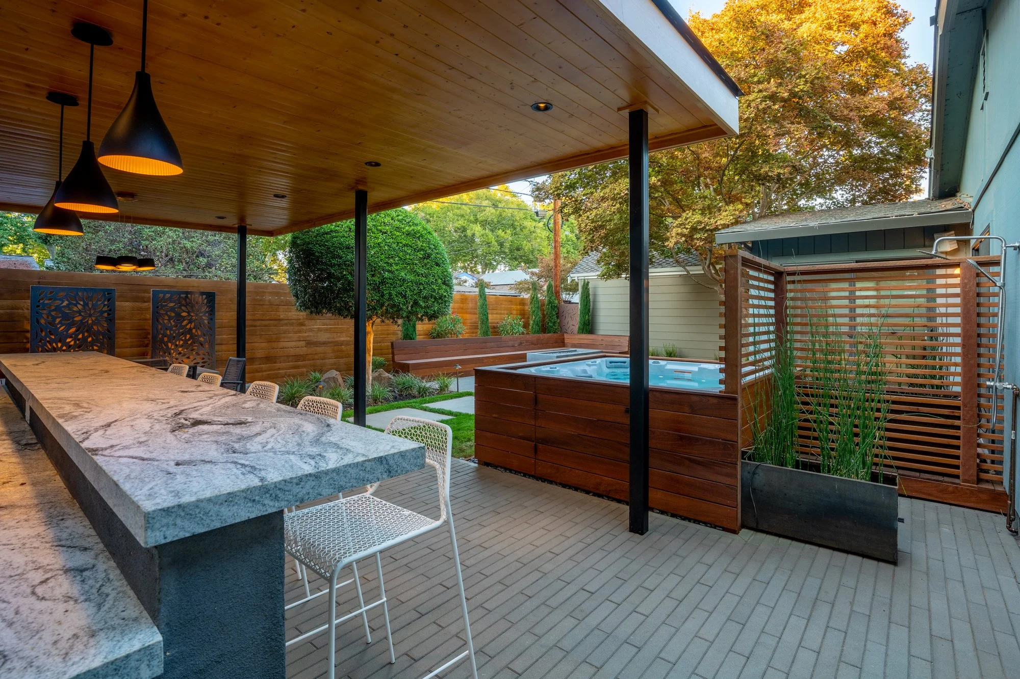 Modern outdoor patio with covered kitchen bar, pendant lights, dining stools, landscaping, and a hot tub behind a privacy screen