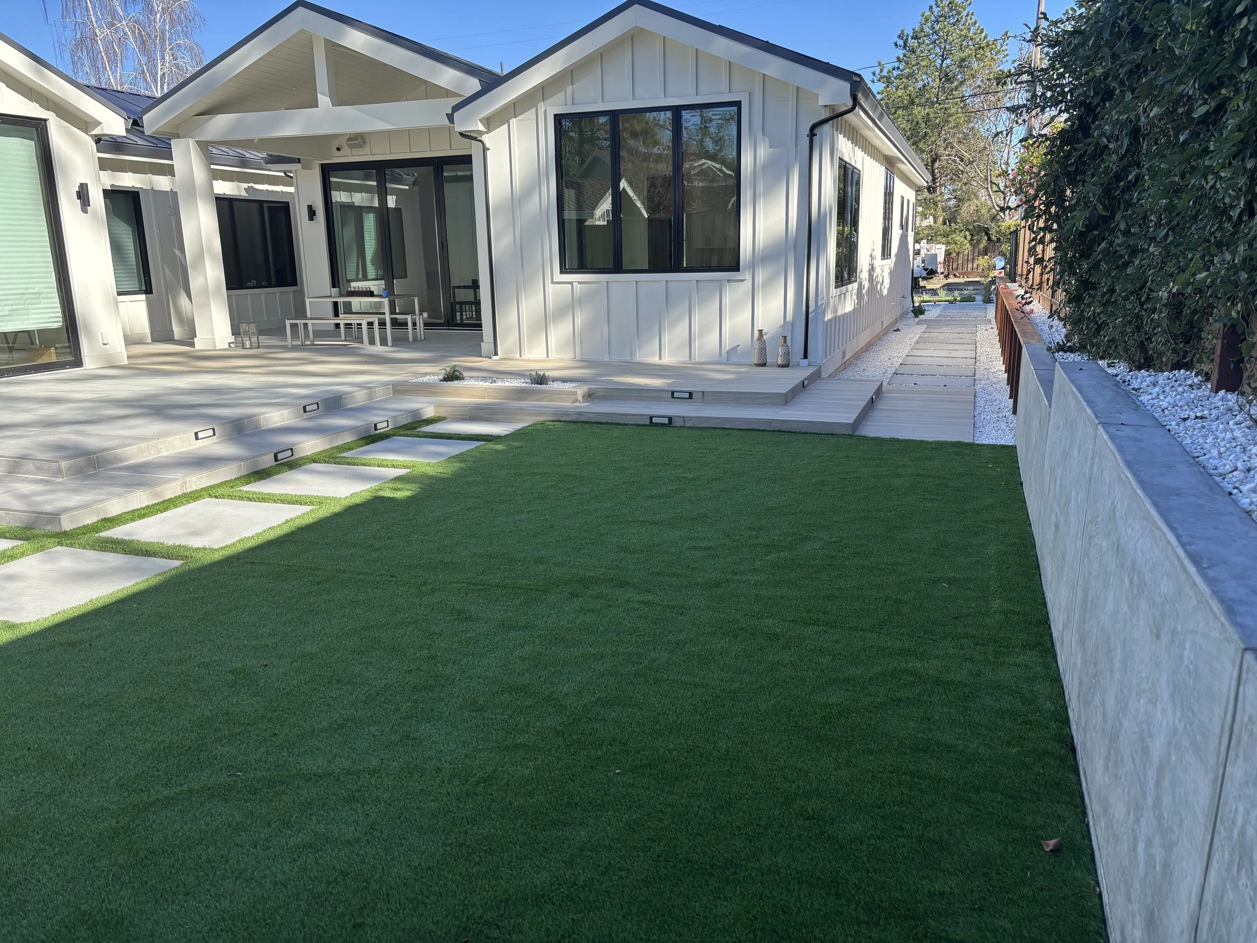 Modern white home with a covered patio, large black-framed windows, landscaped artificial lawn, concrete stepping stones, and wood decking.