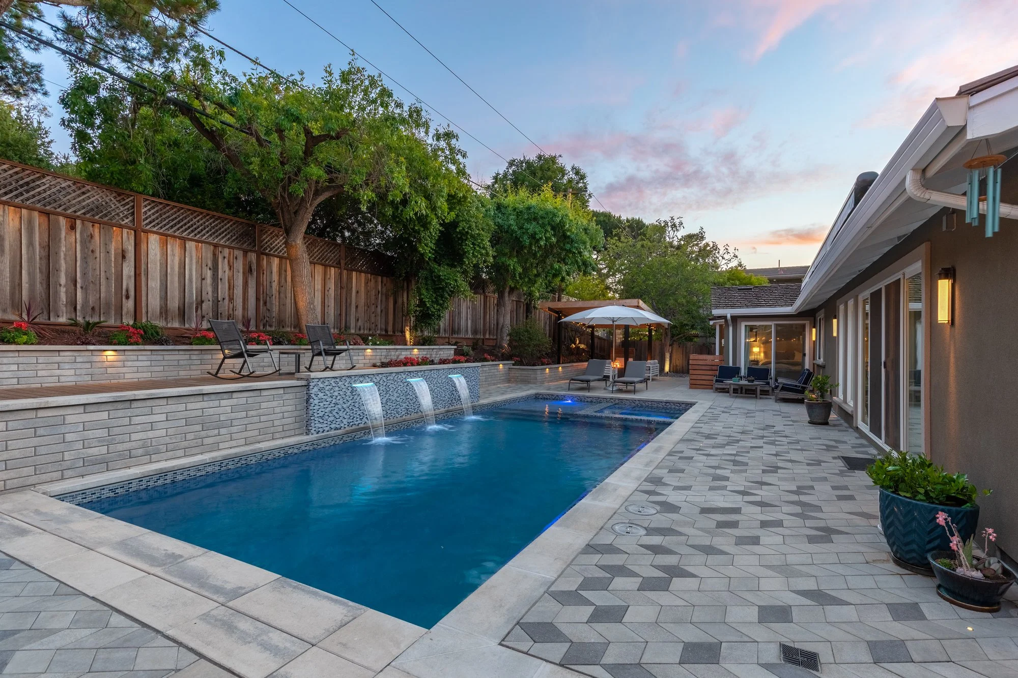 Landscaped backyard with a rectangular swimming pool, cascading water features, lounge chairs, patio seating, and a covered outdoor dining area at sunset.