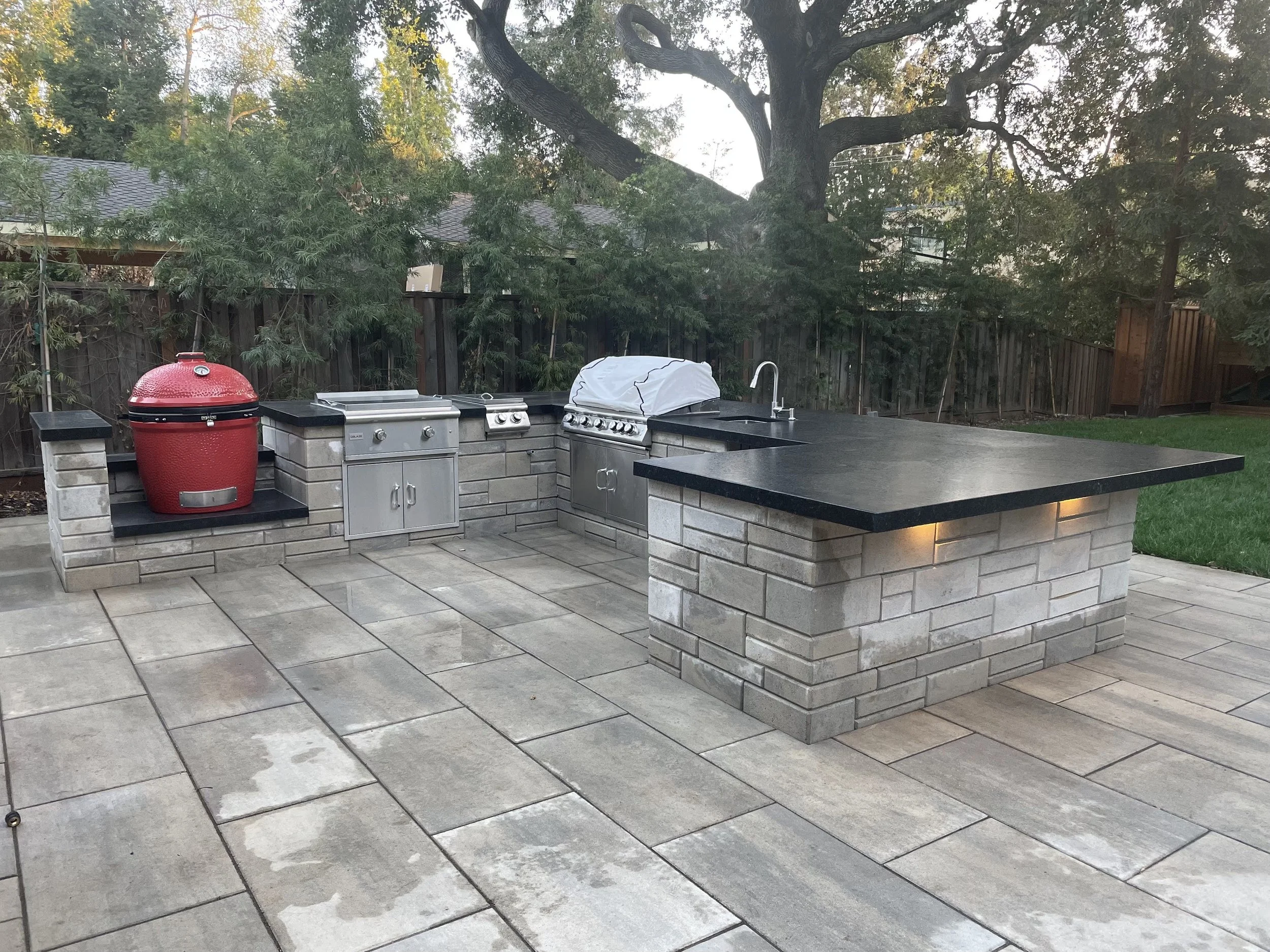 Luxury outdoor kitchen with multiple grills, red kamado smoker, bar seating, stone countertops, and sink in a landscaped backyard