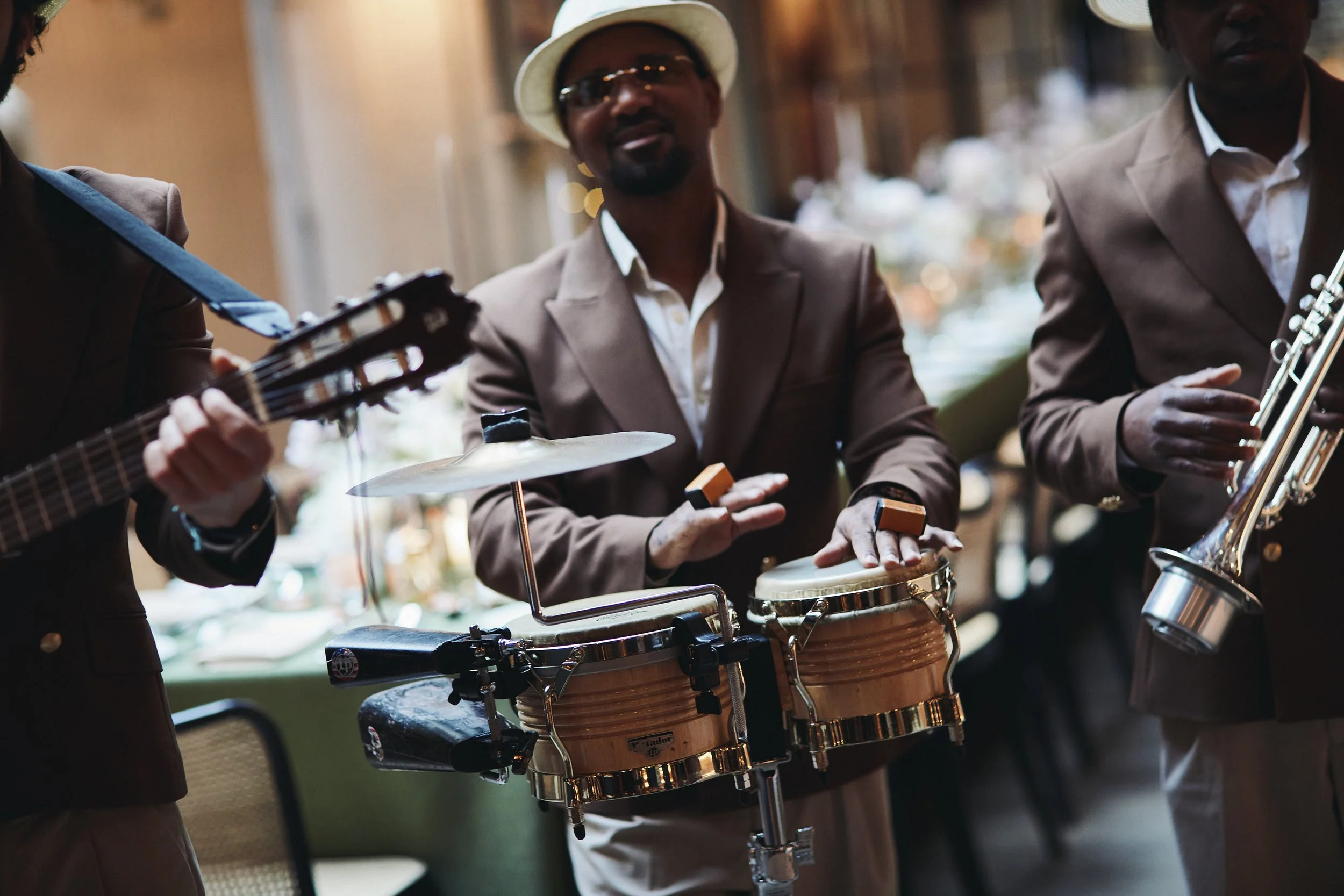 A group of musicians playing instruments at an indoor event. The focus is on a man playing conga drums, wearing a tan suit, white shirt, and a tan hat. Another person is playing a guitar, and a third is playing a saxophone. The background is blurred 