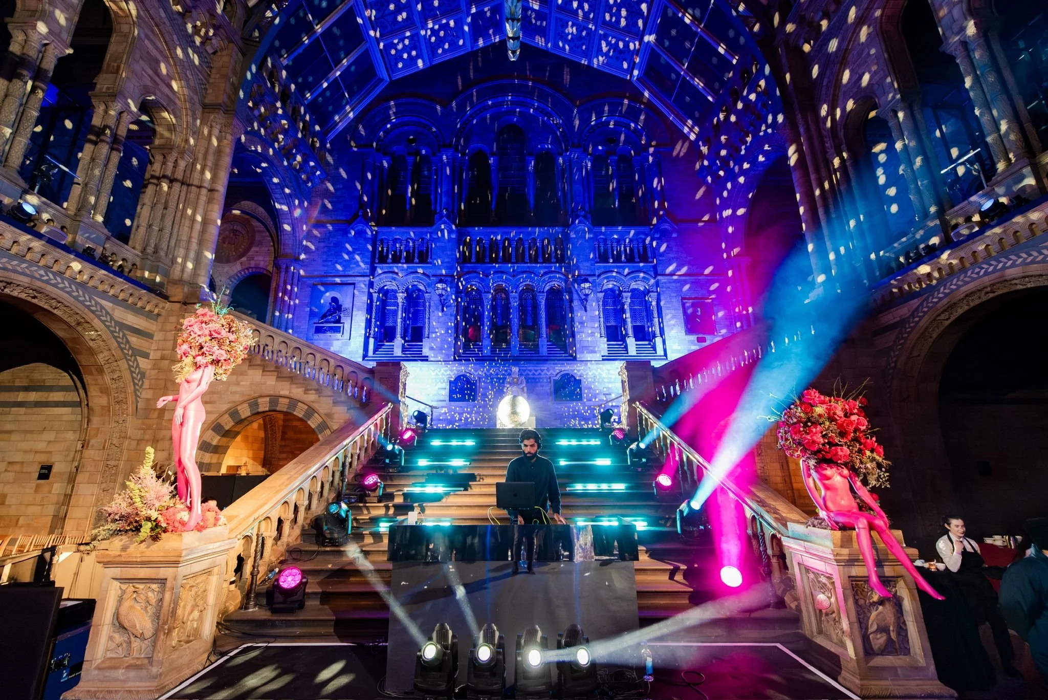 A DJ performs on a stage at the base of a grand staircase inside a large, ornate hall with arched architecture, dramatic colorful lighting, and floral decorations on either side of the steps.