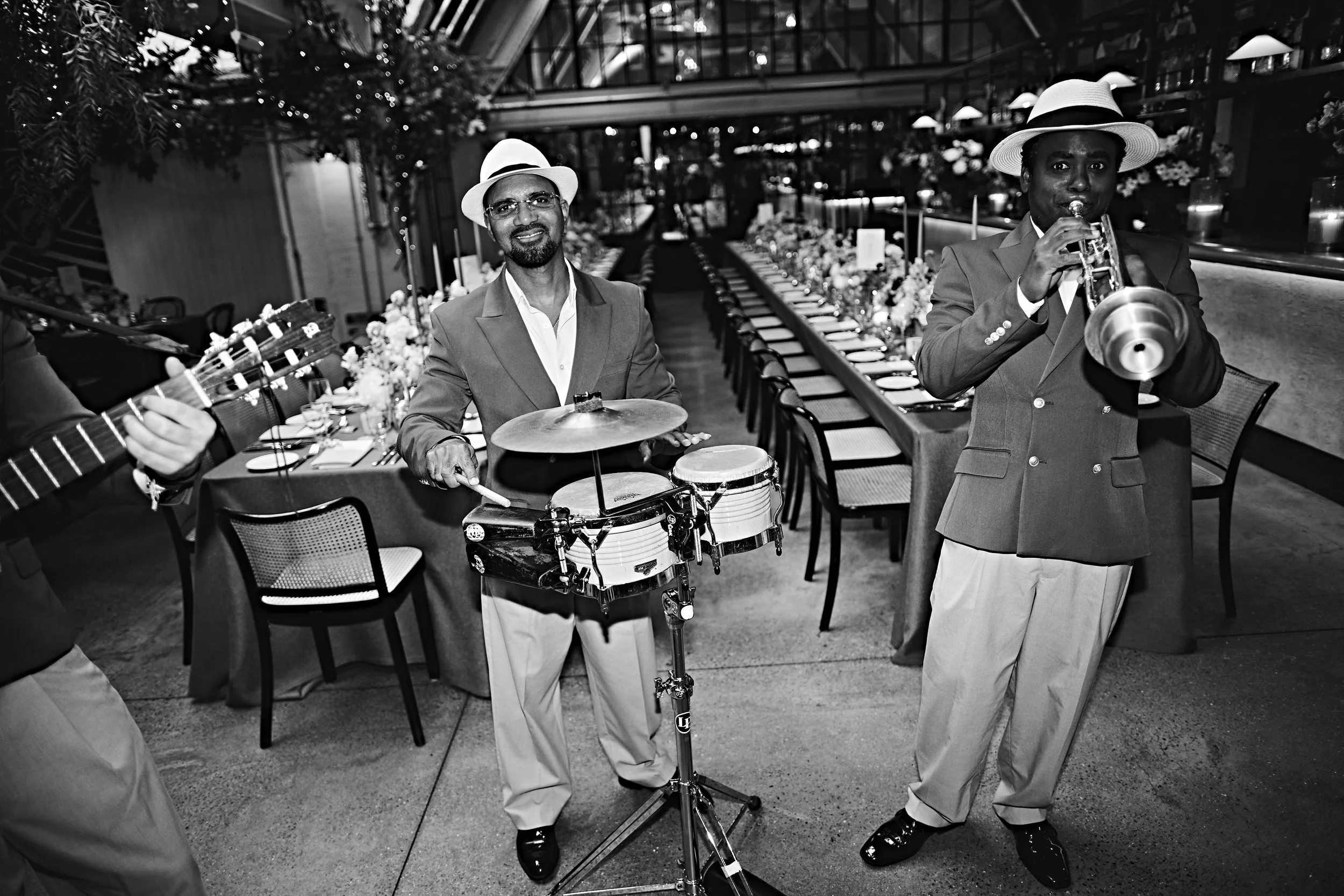 Black and white photo of a man playing drums and a man playing trumpet at a celebration or event in a modern indoor venue with a long dining table decorated with flowers.