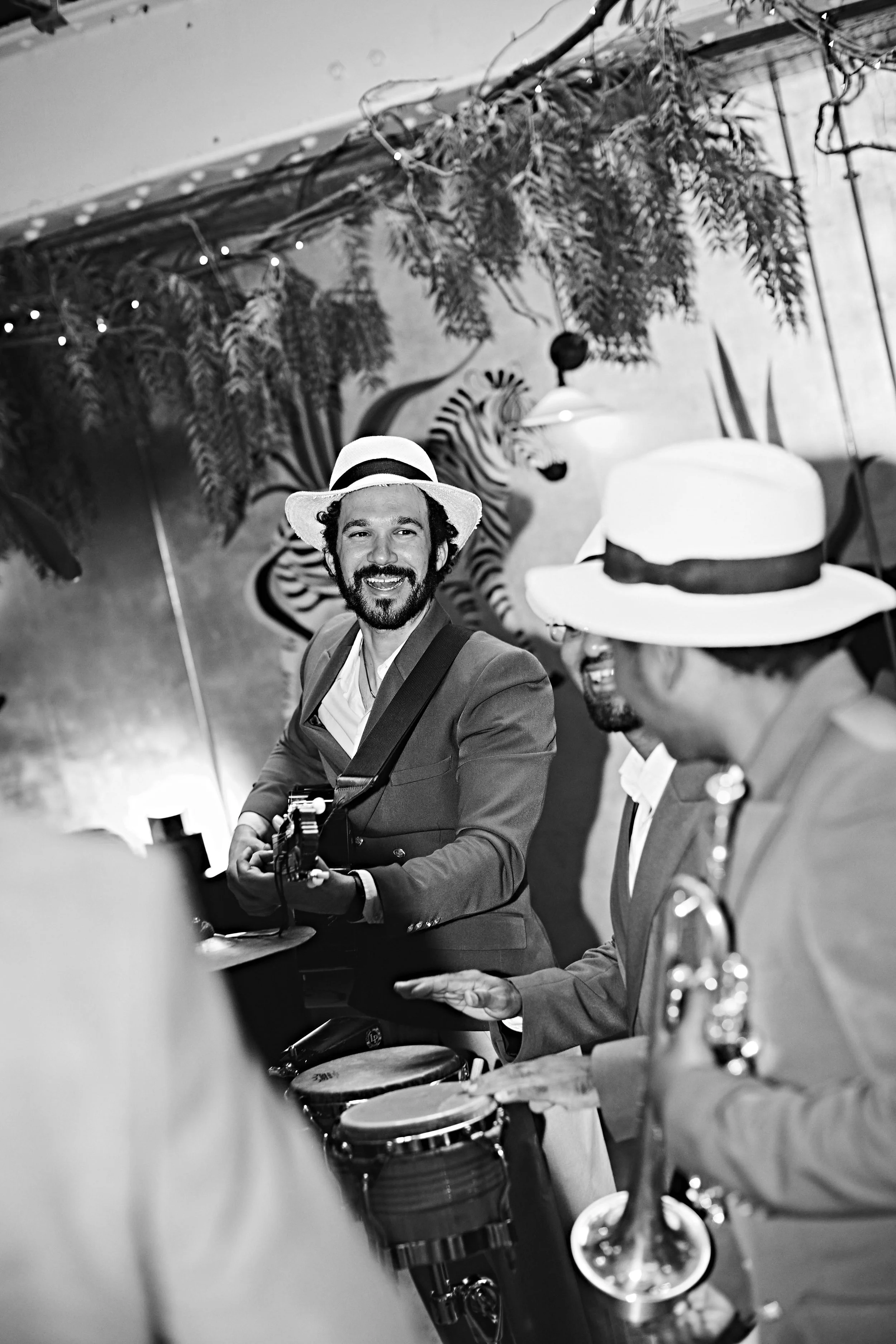 A group of people, including a man in a suit and a hat, playing musical instruments such as drums and a saxophone in a lively indoor setting decorated with plants and tiger mural.