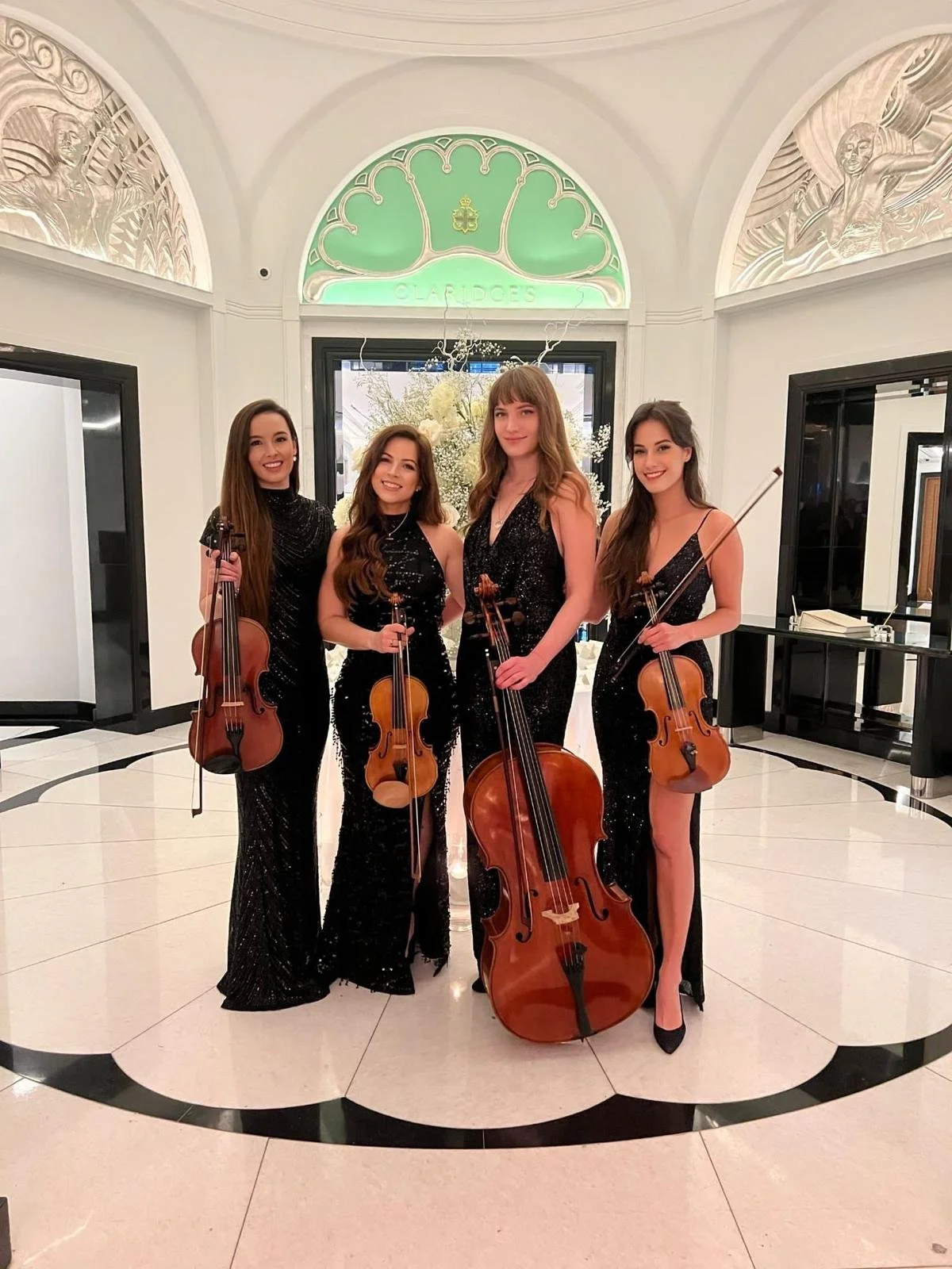 Four women musicians dressed in elegant black gowns pose in a grand lobby with white marble floors and ornate decor. Three hold violins and one holds a cello, standing beneath an arched design with the word 'Claridges' illuminated in green above them