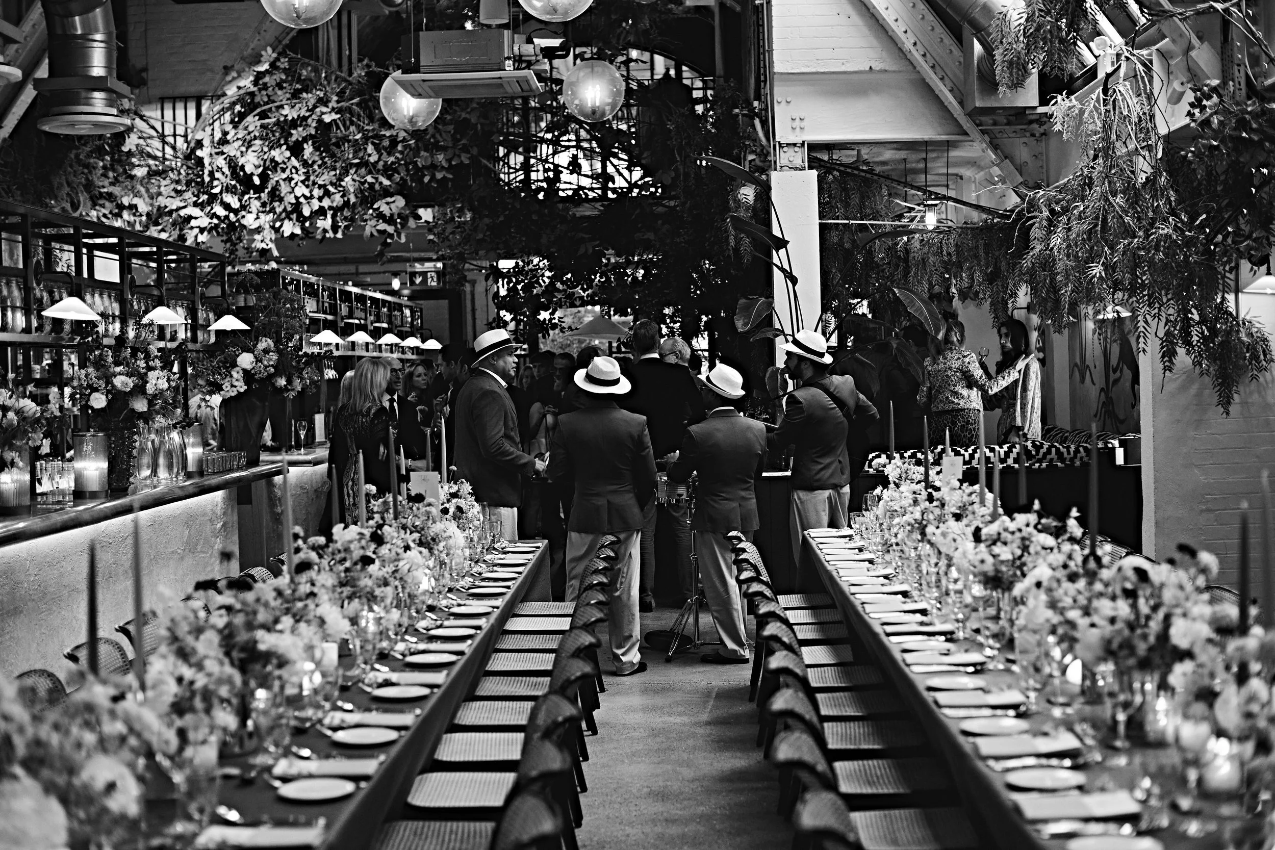 Black and white photo of a decorated event space with long tables set for a meal, surrounded by people dressed in formal attire, some wearing hats, with floral arrangements and hanging lights.