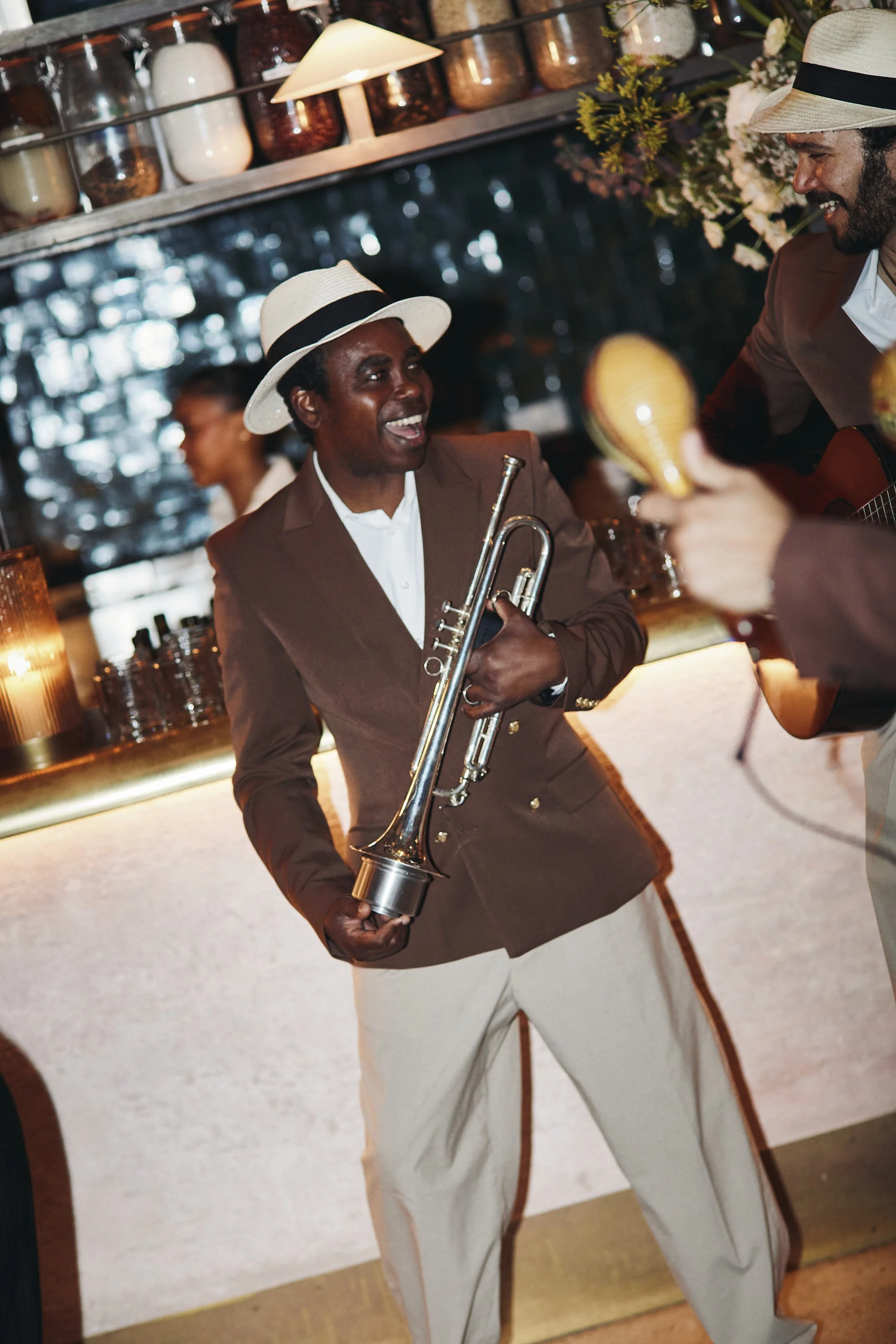 A man wearing a brown suit, white shirt, and a white hat with a black band, smiling and holding a silver trumpet in a bar or restaurant setting with decorative jars on shelves and floral arrangements in the background.