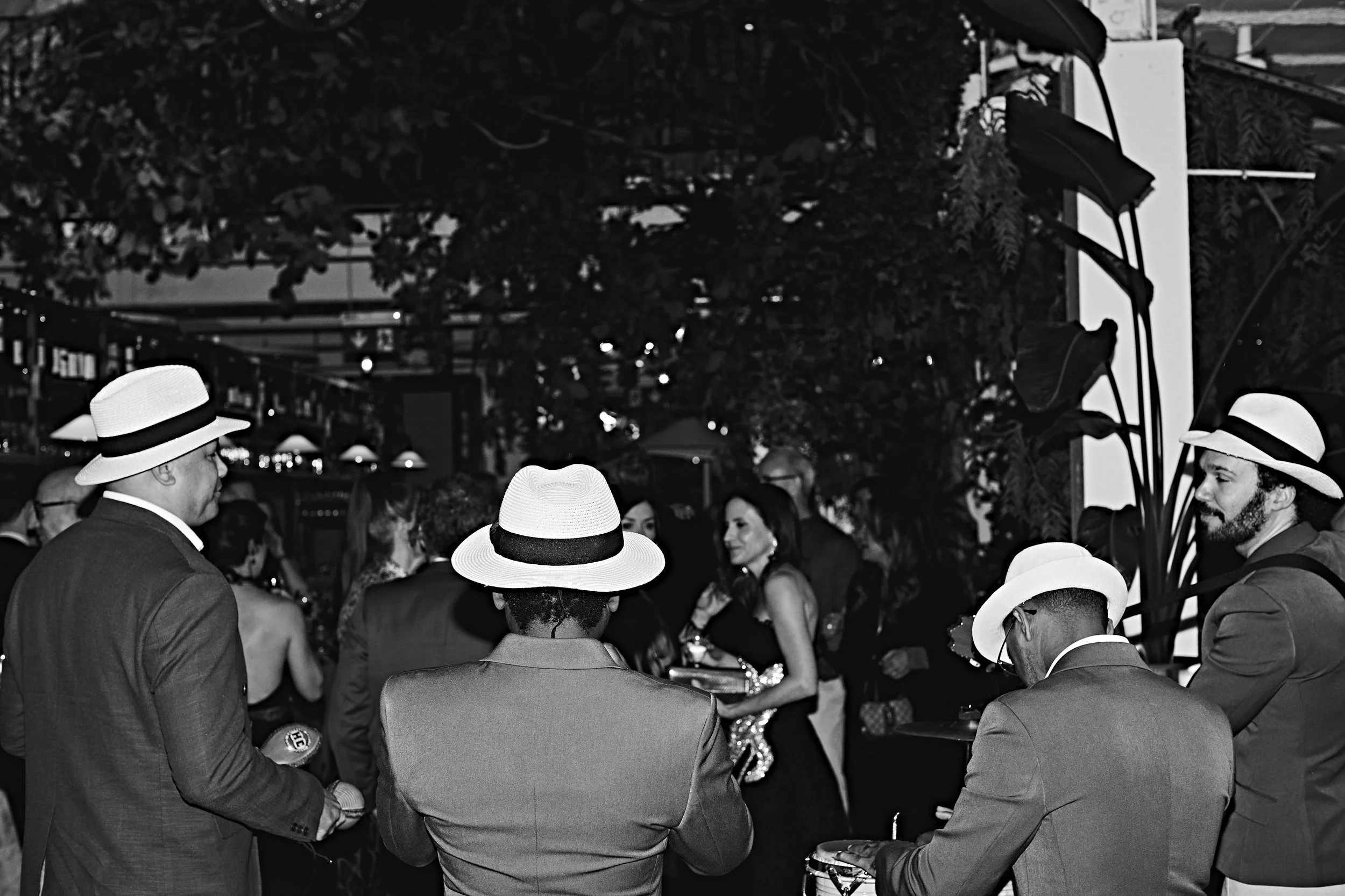 A black-and-white photo of a social gathering. Several people wearing formal attire and hats are engaged in conversation outdoors, with greenery and some structures visible in the background.