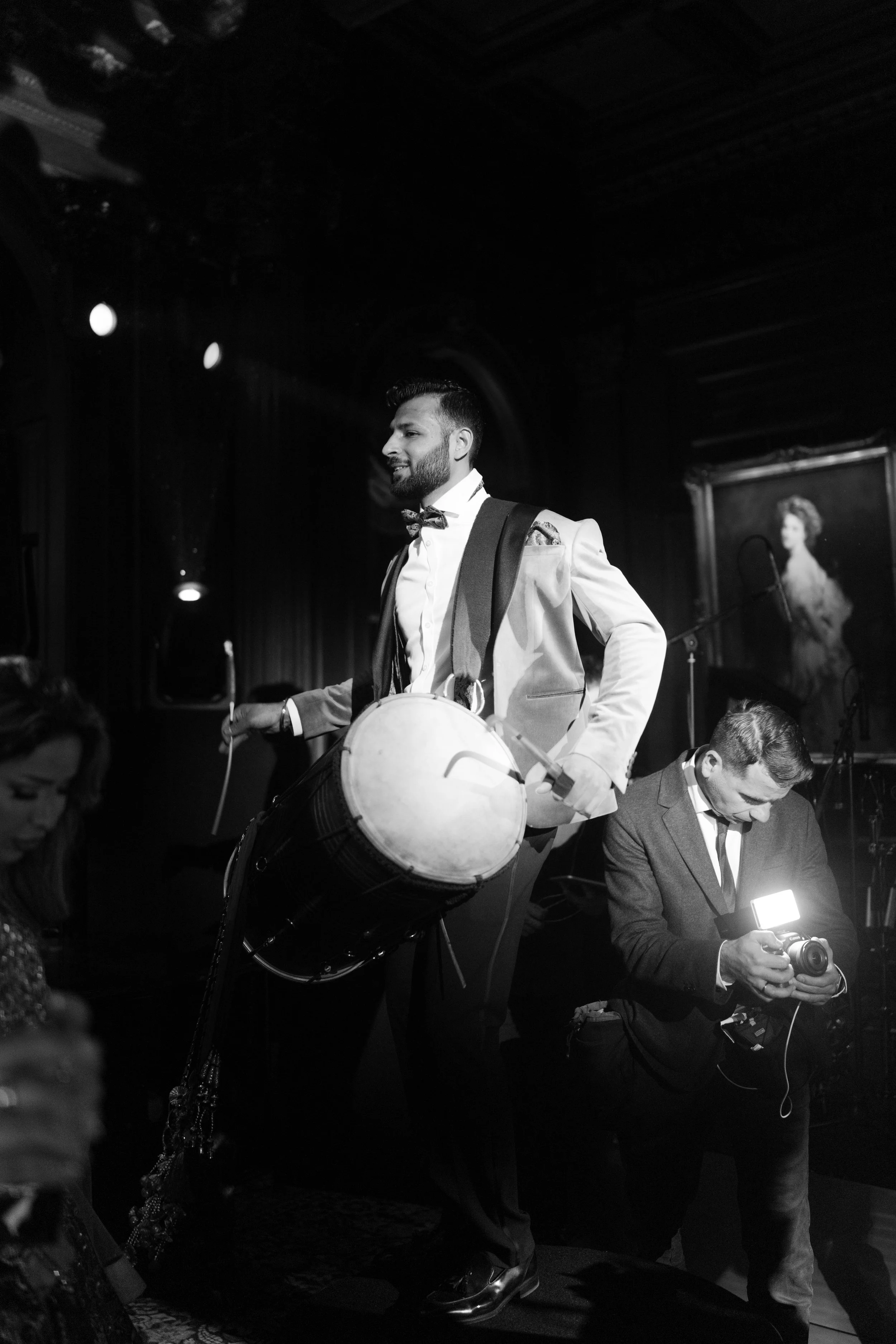 At the center, a well-dressed man is playing a large drum, wearing a suit with a bow tie, which gives a formal yet celebratory vibe. His posture and expression suggest he’s actively entertaining the crowd. To his right, another man in a suit is focus