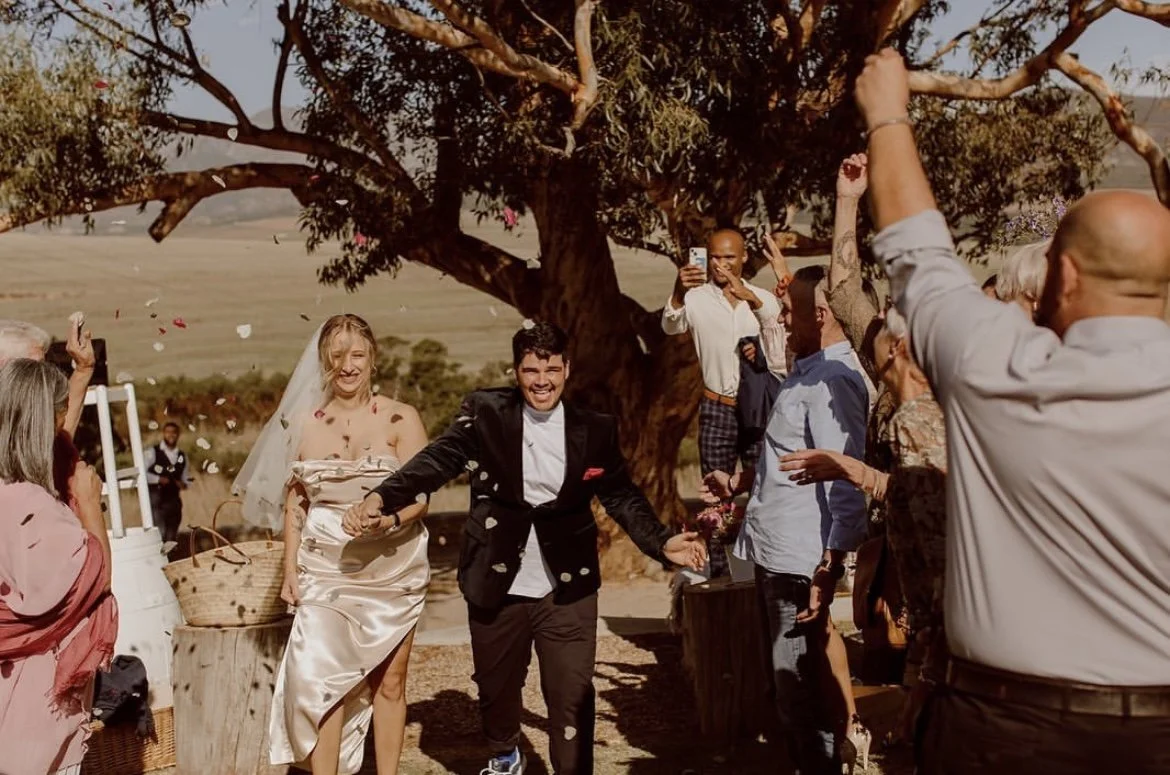 A wedding celebration outdoors with a wedding couple walking hand in hand through a shower of flower petals, surrounded by guests cheering and taking photos under a large tree.