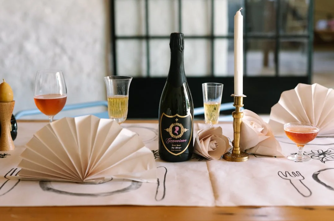 Table setting with champagne, wine, and cocktail glasses, a bottle of champagne, a candle, a folded napkin, and decorative roses.