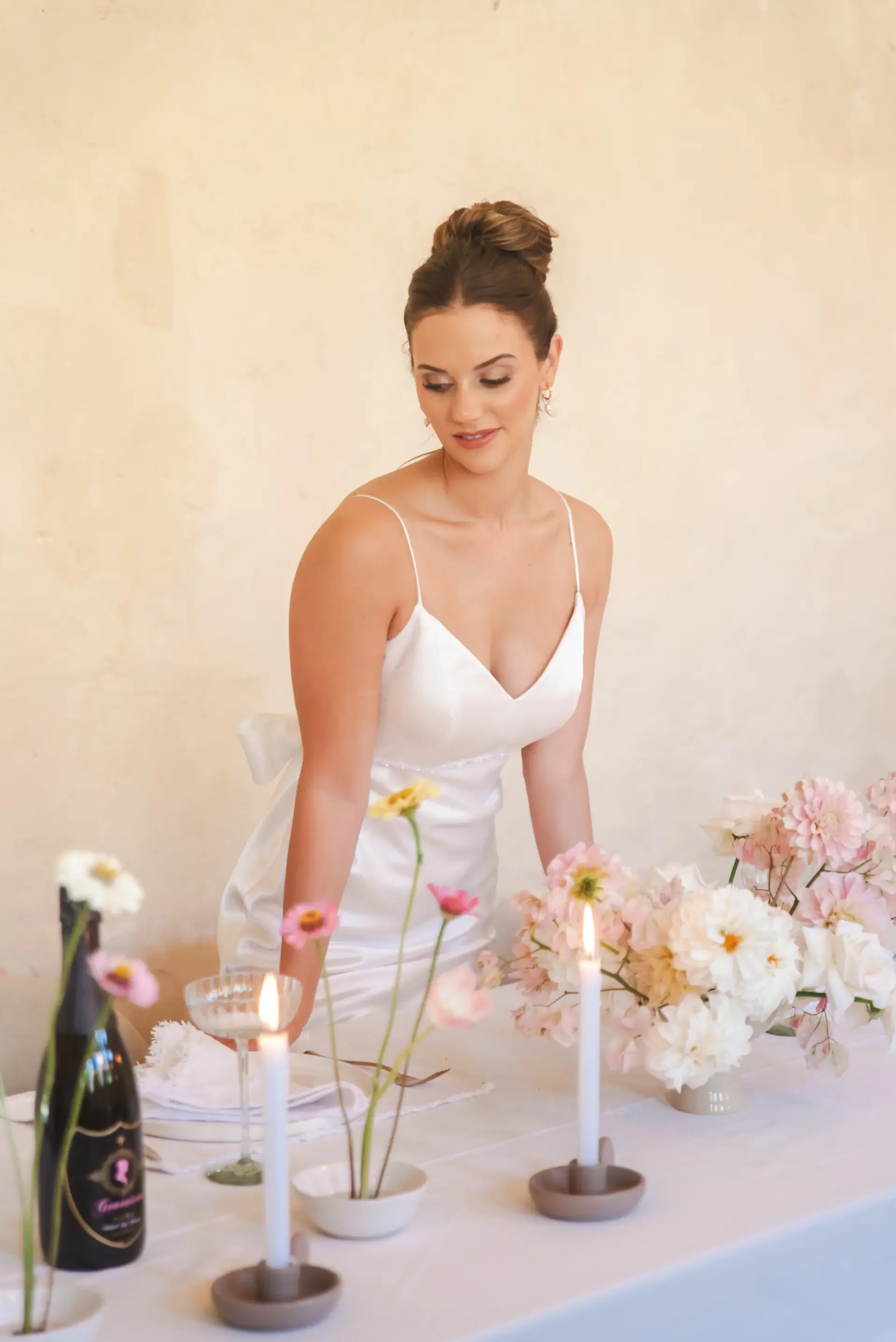 A woman in a white dress standing behind a table decorated with pink and white flowers, lit candles, and a bottle of champagne, with a beige wall in the background.