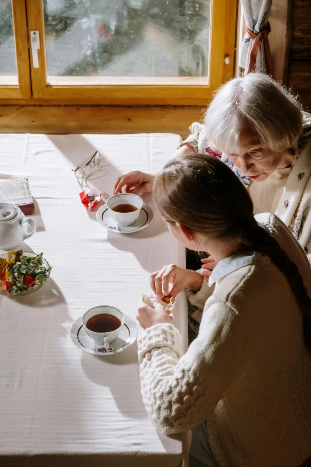 Grandmother and granddaughter having tea at a kitchen table, sharing stories and family history questions in warm afternoon light.