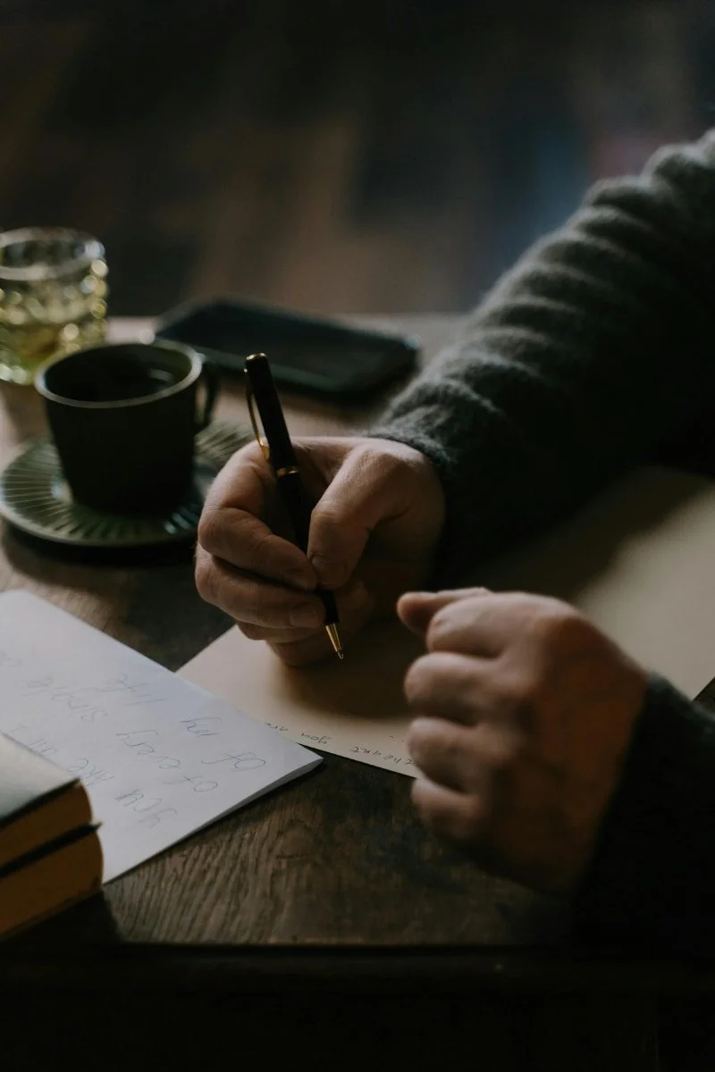 Hands writing notes at a table with a cup of coffee, a notebook, and soft light in the background.