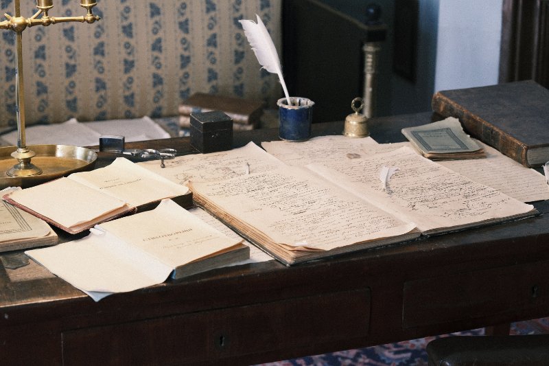 An antique writing desk covered with handwritten documents, books, an inkpot, and a quill shows the kinds of historical records researchers may encounter when using AI for genealogy research to plan their next searches.