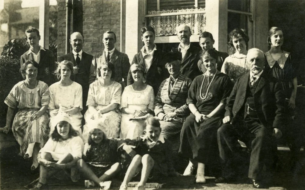 Black-and-white portrait of a large family group posed outside a house, showing multiple generations and extended relatives together.