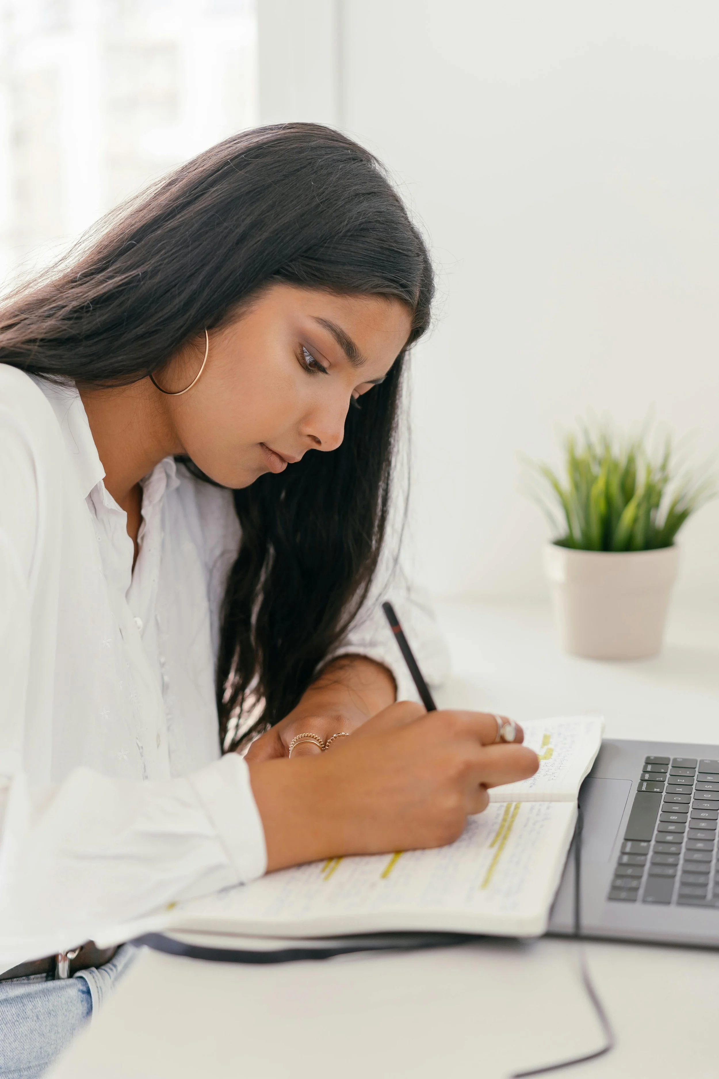 Woman writing in a notebook, focused and thoughtful, with a laptop and plant nearby—perfectly capturing the modern family historian organizing her genealogy workbook.