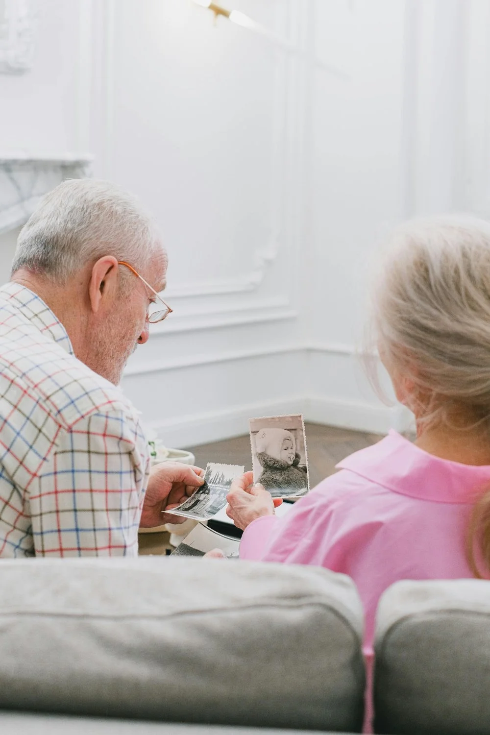 Elderly couple sitting on a couch looking at old photographs, sparking family history questions about their childhood and ancestors.