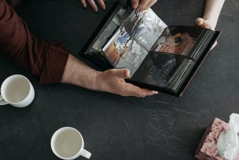 Two people sit at a dark table, flipping through a photo album together. Cups and tissues nearby hint at shared memories. A quiet moment of preserving family stories through old photos.