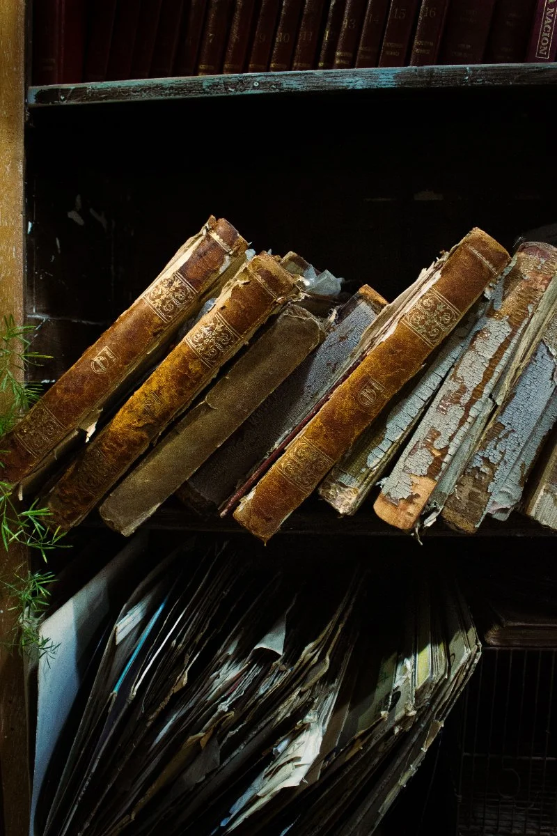 A collection of aging books and records sits on a shelf, highlighting the types of historical sources genealogists use to answer family history questions. Valuable information is often hidden in archives, libraries, and local record collections.