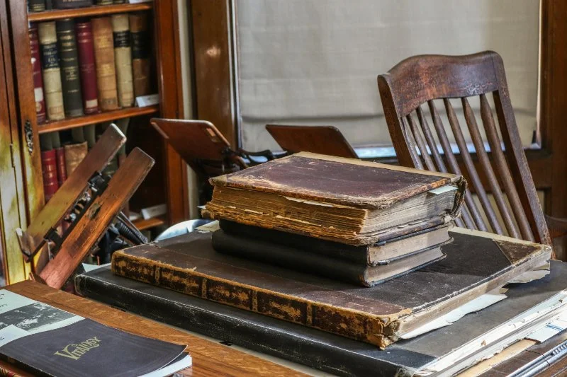 Worn antique books and oversized historical volumes rest on a wooden table inside an old research library | AI and genealogy.