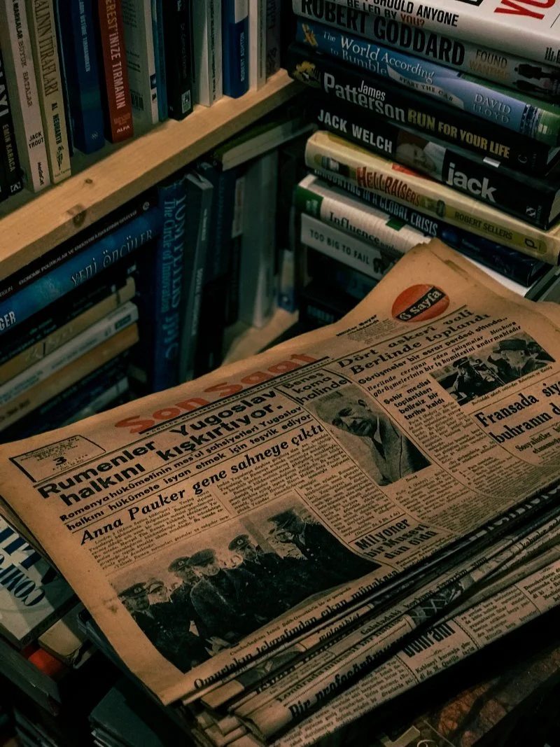Stack of vintage genealogy newspapers resting among books in a home library, representing historical newspaper archives for ancestor research.